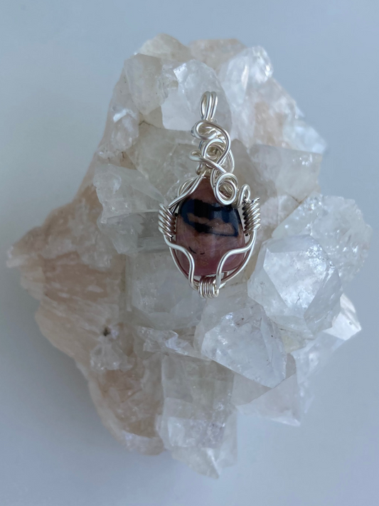 Rhodonite Pendant – Front view of a wire-wrapped pink and black rhodonite stone pendant, handcrafted in silver-plated copper wire with a unique spiral design — photographed on a clear apophyllite crystal cluster with a white background.