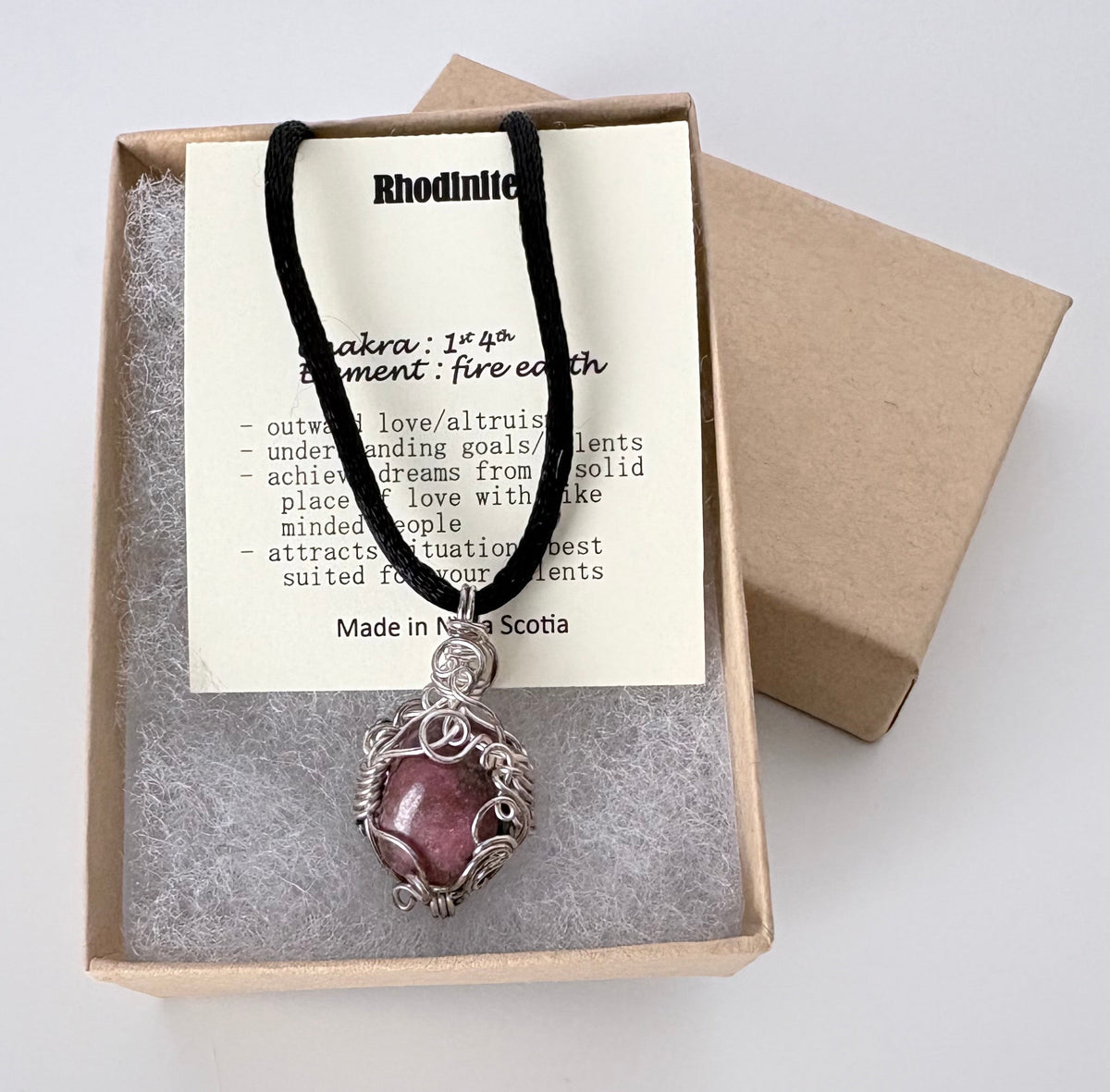 Necklace with a silver wire wrapped rhodonite pendant on an information card with text explaining the folklore and metaphysical properties placed in a brown paper kraft jewelry box for gift giving all on a white surface.