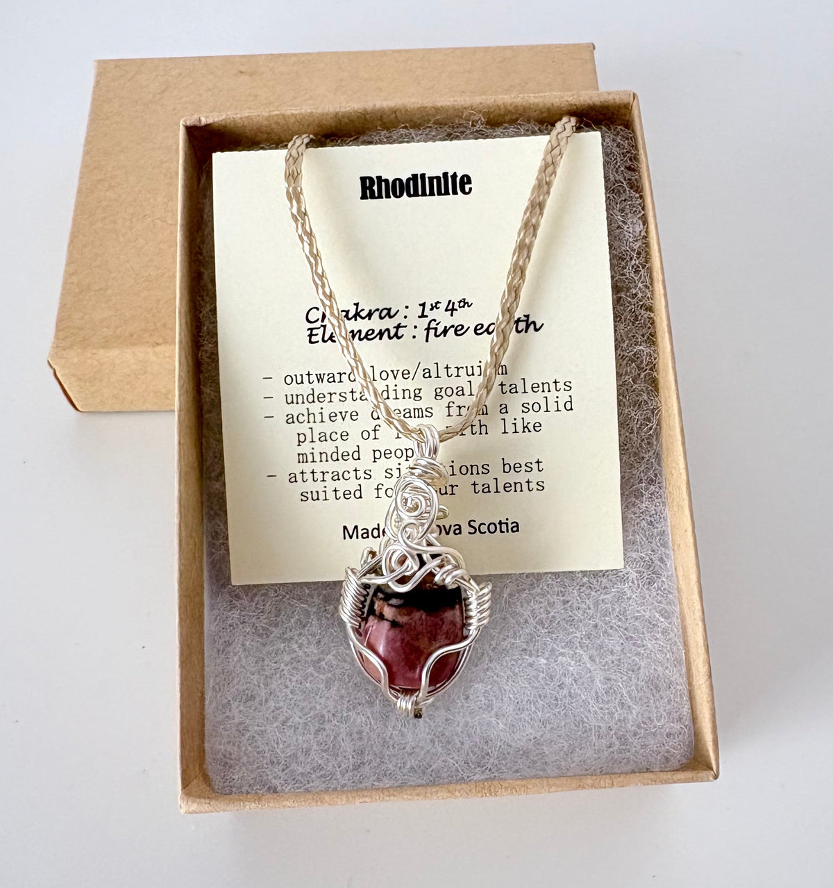 Necklace with a silver wire wrapped rhodonite pendant on an information card with text explaining the folklore and metaphysical properties placed in a brown paper kraft jewelry box for gift giving all on a white surface.