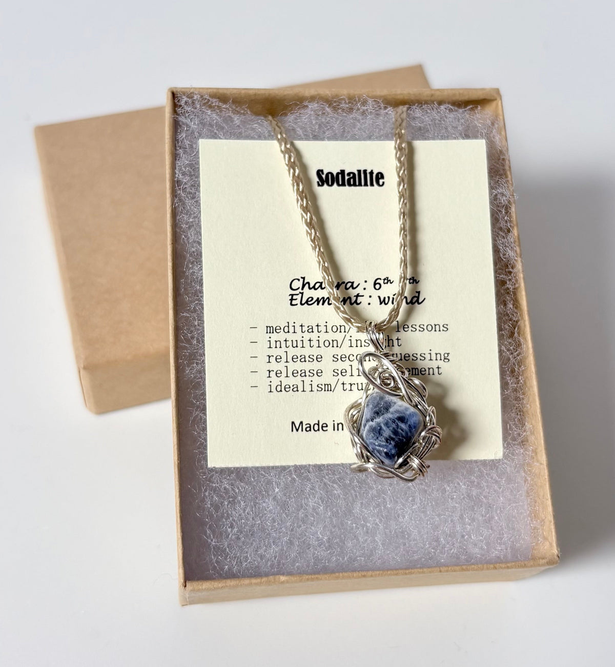 Necklace with a silver wire wrapped sodalite pendant on an information card with text explaining the folklore and metaphysical properties placed in a brown paper kraft jewelry box for gift giving all on a white surface.