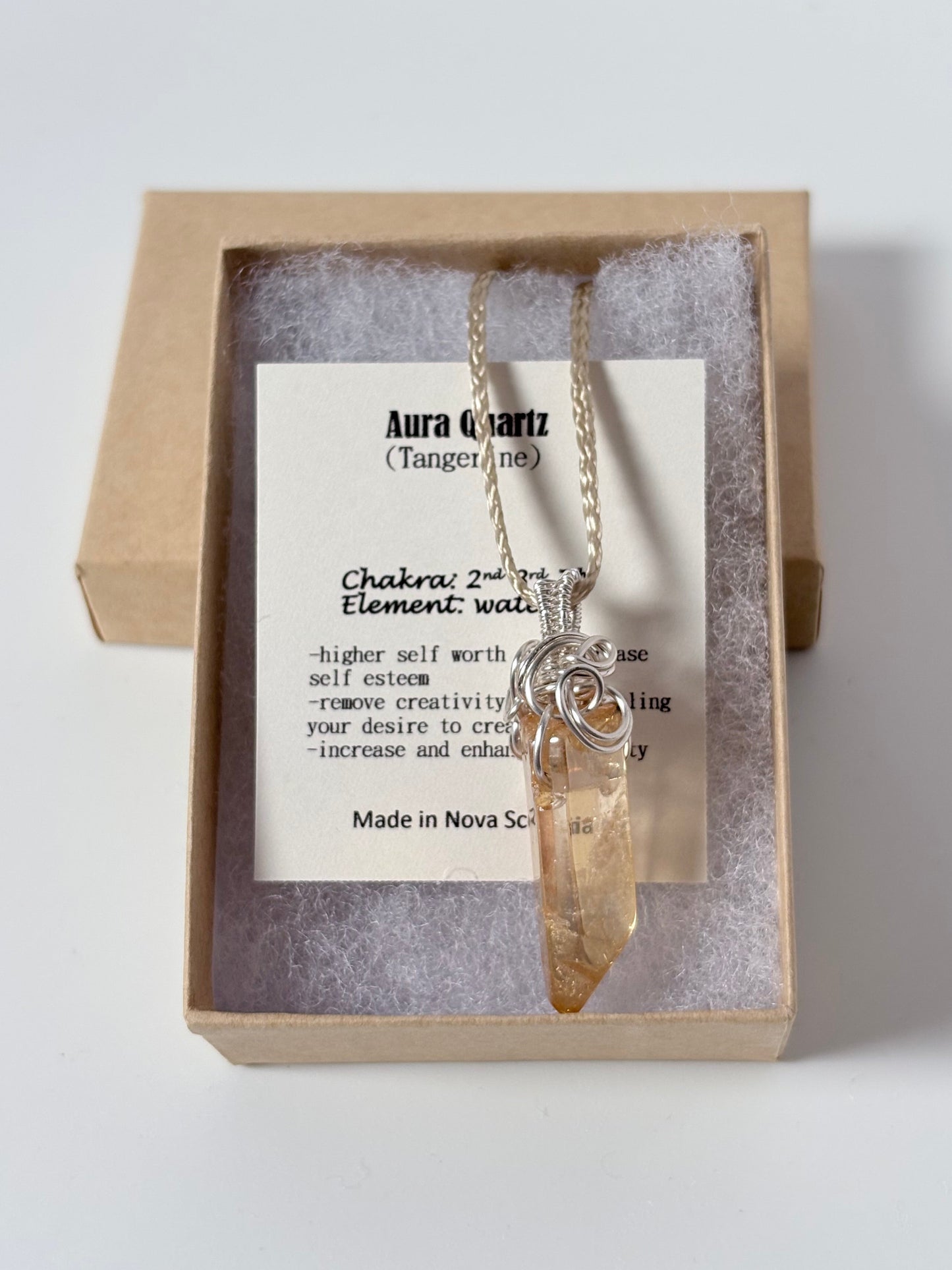 Necklace with a silver wire wrapped tangerine aura Quartz pendant on an information card with text explaining the folklore and metaphysical properties placed in a brown paper kraft jewelry box for gift giving all on a white surface.