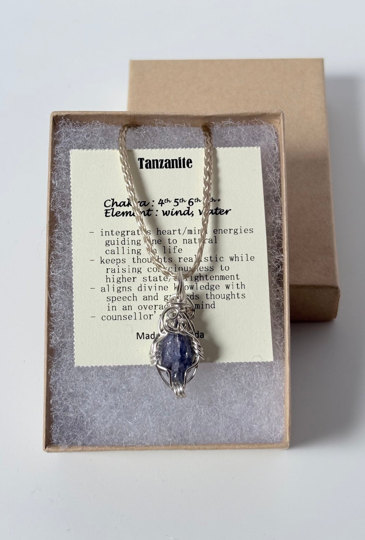 Necklace with a silver wire wrapped tanzanite pendant on an information card with text explaining the folklore and metaphysical properties placed in a brown paper kraft jewelry box for gift giving all on a white surface.