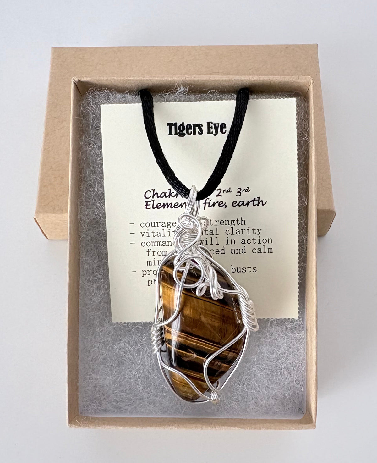 Necklace with a silver wire wrapped tigers eye pendant on an information card with text explaining the folklore and metaphysical properties placed in a brown paper kraft jewelry box for gift giving all on a white surface.