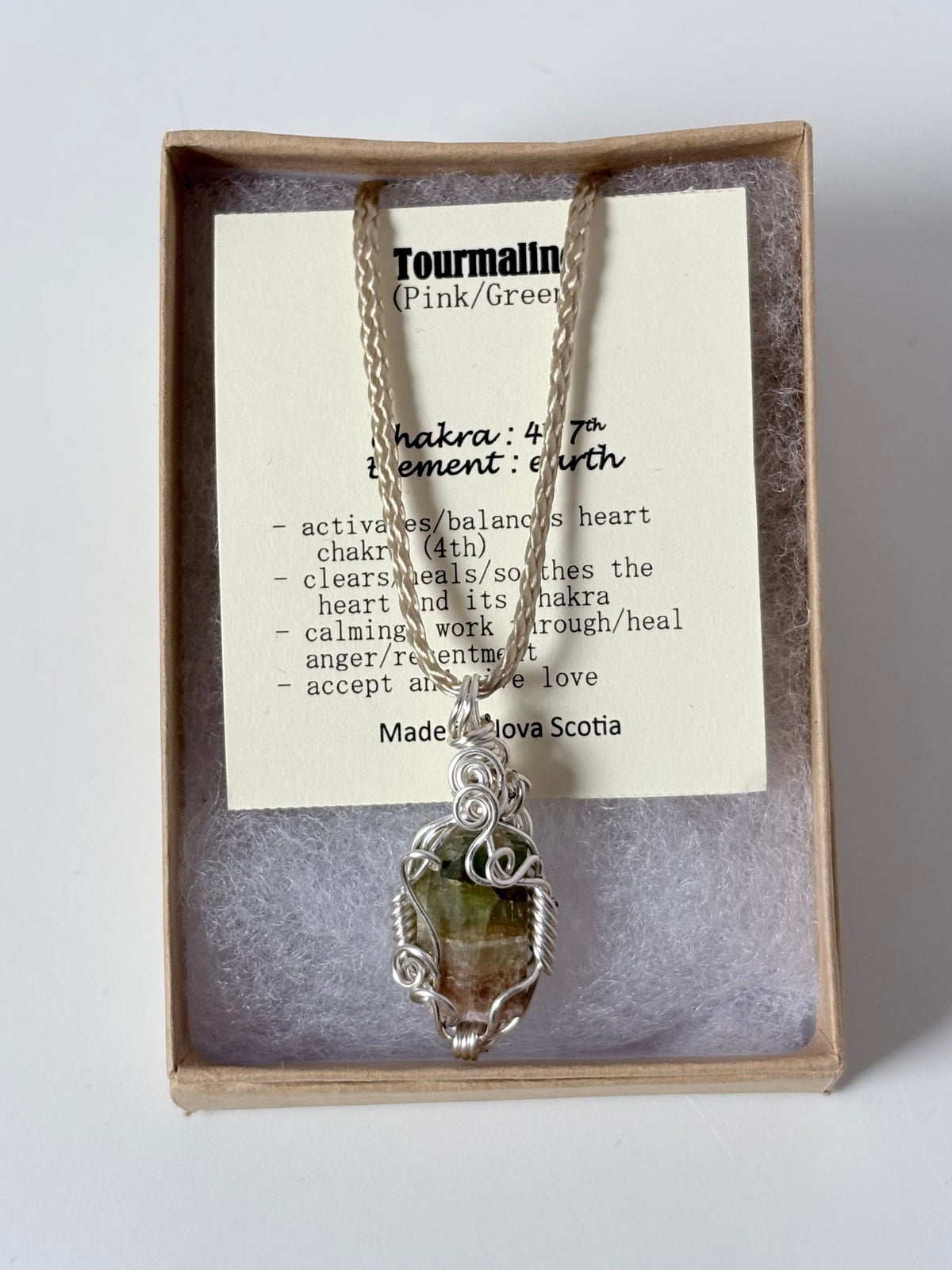 Necklace with a silver wire wrapped tourmaline (pink/green) pendant on an information card with text explaining the folklore and metaphysical properties placed in a brown paper kraft jewelry box for gift giving all on a white surface.
