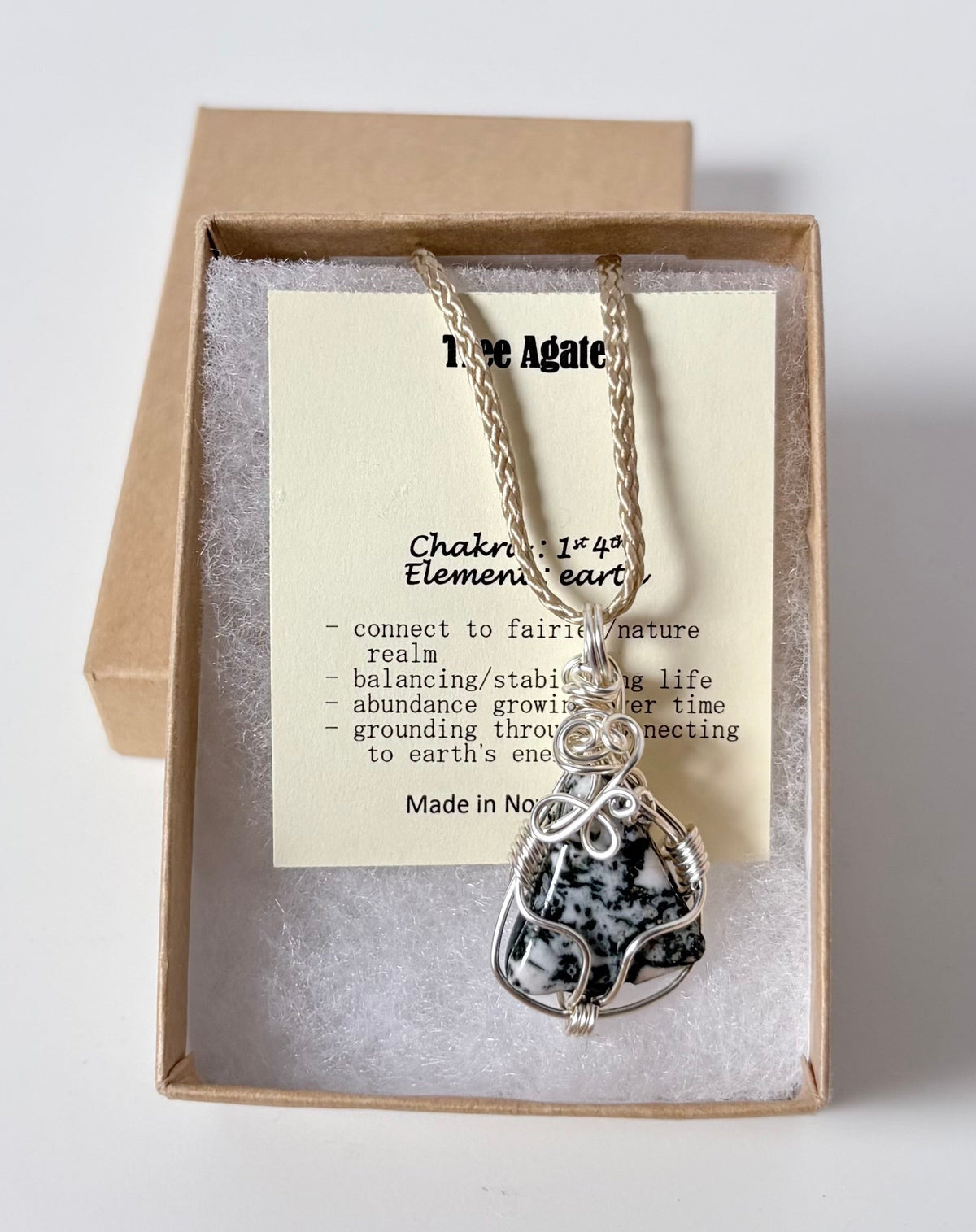 Necklace with a silver wire wrapped tree agate pendant on an information card with text explaining the folklore and metaphysical properties placed in a brown paper kraft jewelry box for gift giving all on a white surface.