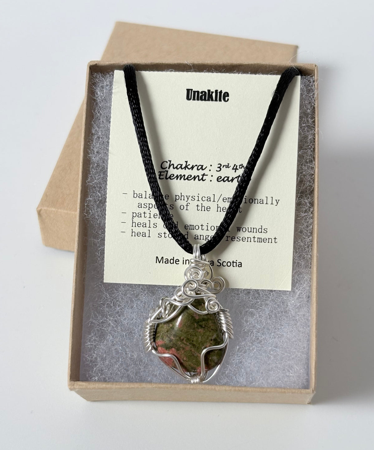 Necklace with a silver wire wrapped unakite pendant on an information card with text explaining the folklore and metaphysical properties placed in a brown paper kraft jewelry box for gift giving all on a white surface.