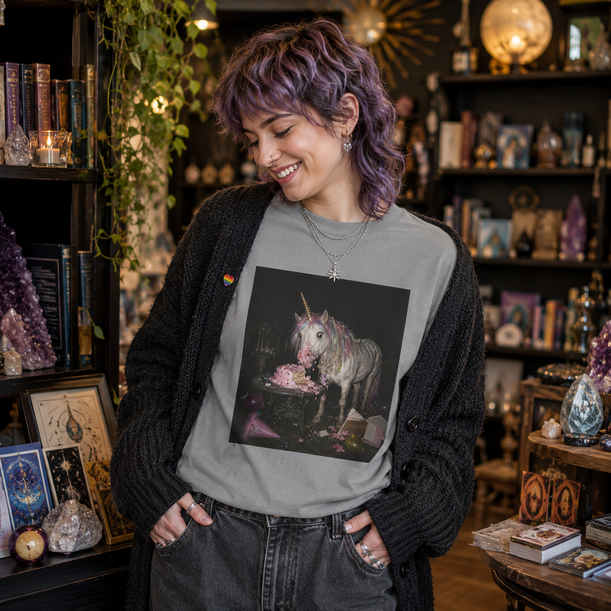 Grey graphic t-shirt with a retro-style photo of a unicorn stealing pink birthday cake in the dark at a party. the shirt is shown on a model in a cozy spiritual shop with crystals, books and warm lighting.