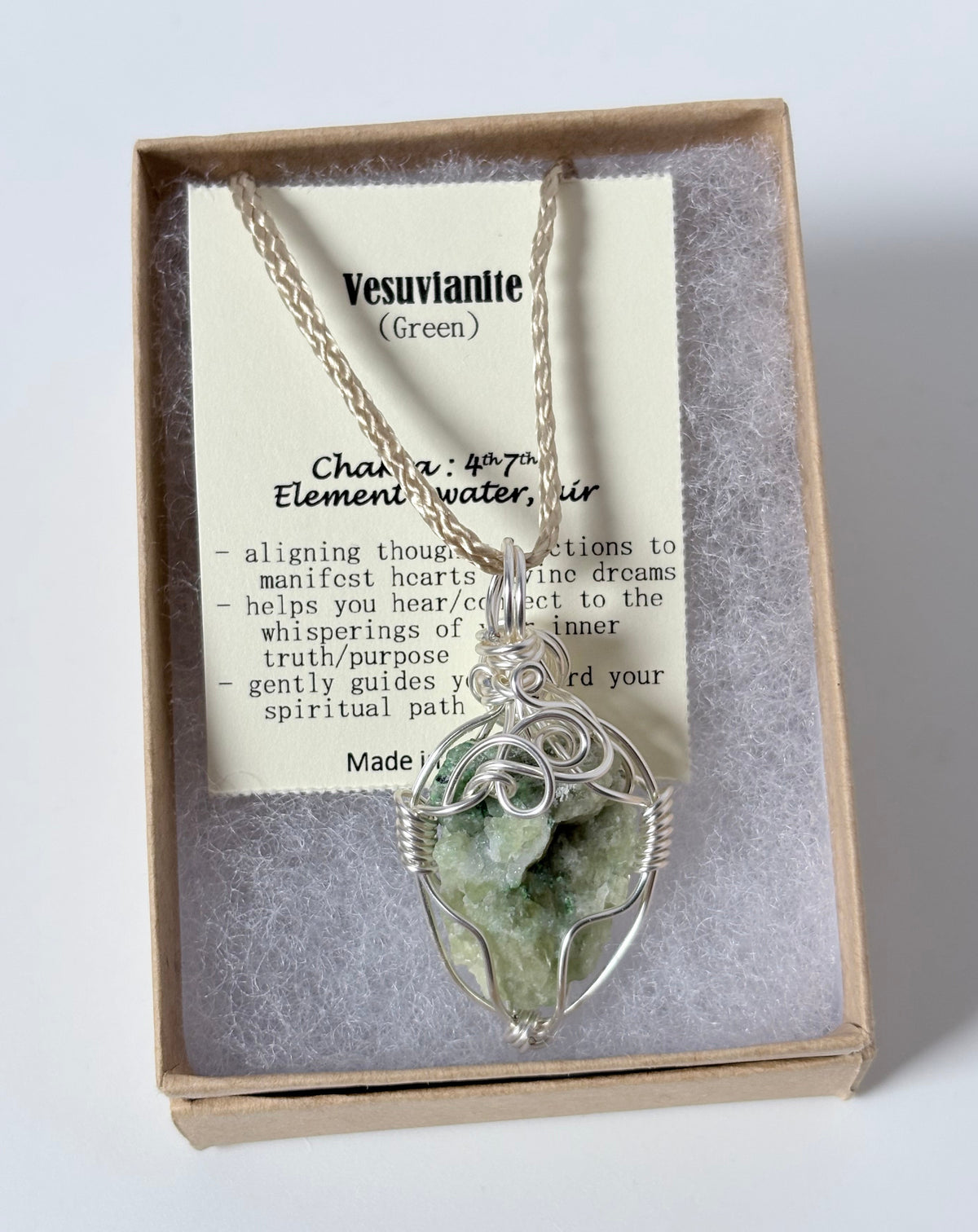 Necklace with a silver wire wrapped vesuvianite pendants pendant on an information card with text explaining the folklore and metaphysical properties placed in a brown paper kraft jewelry box for gift giving all on a white surface.
