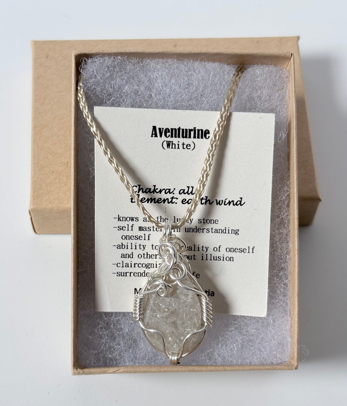Necklace with a silver wire wrapped aventurine (white) pendant on an information card with text explaining the folklore and metaphysical properties placed in a brown paper kraft jewelry box for gift giving all on a white surface.