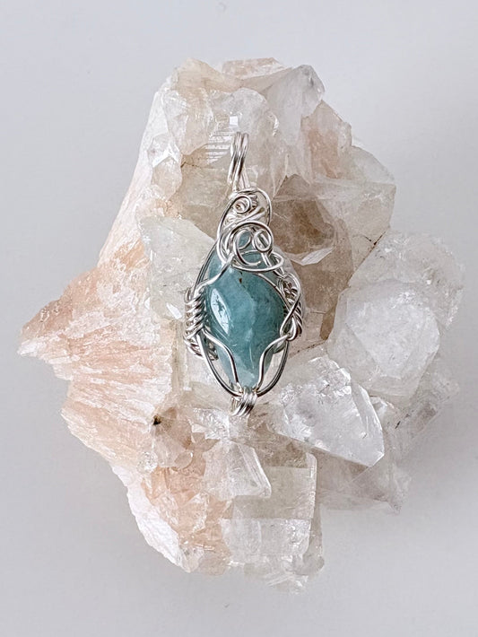 silver wire wrapped aquamarine pendant displayed on a clear apophyllite cluster all on a white surface.