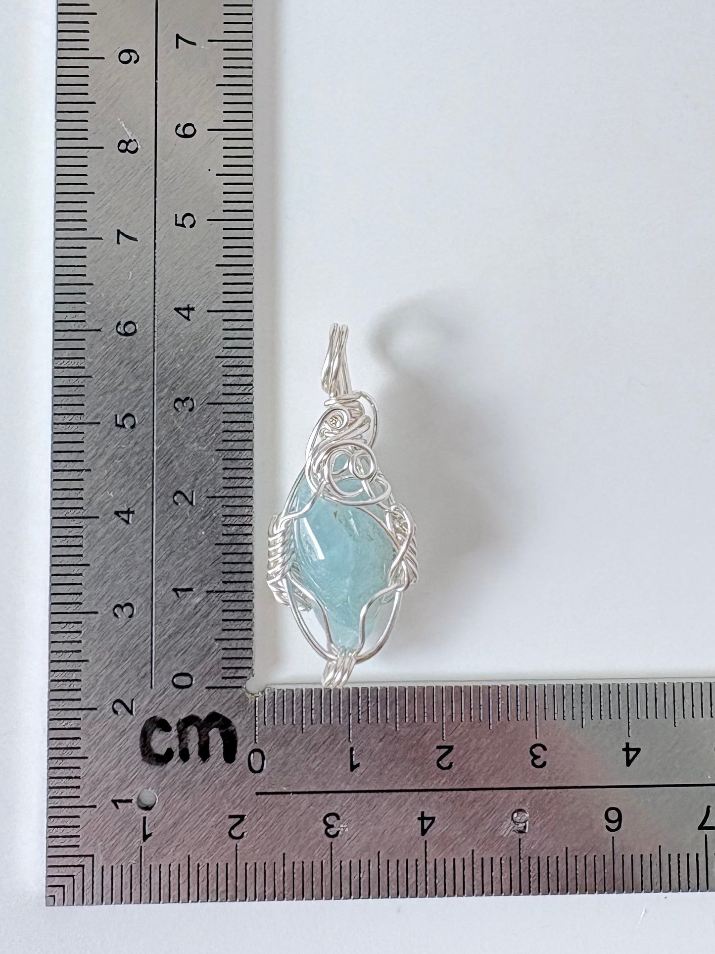 silver wire wrapped aquamarine pendant next to an "L" shaped ruler for scale all against a white background.
