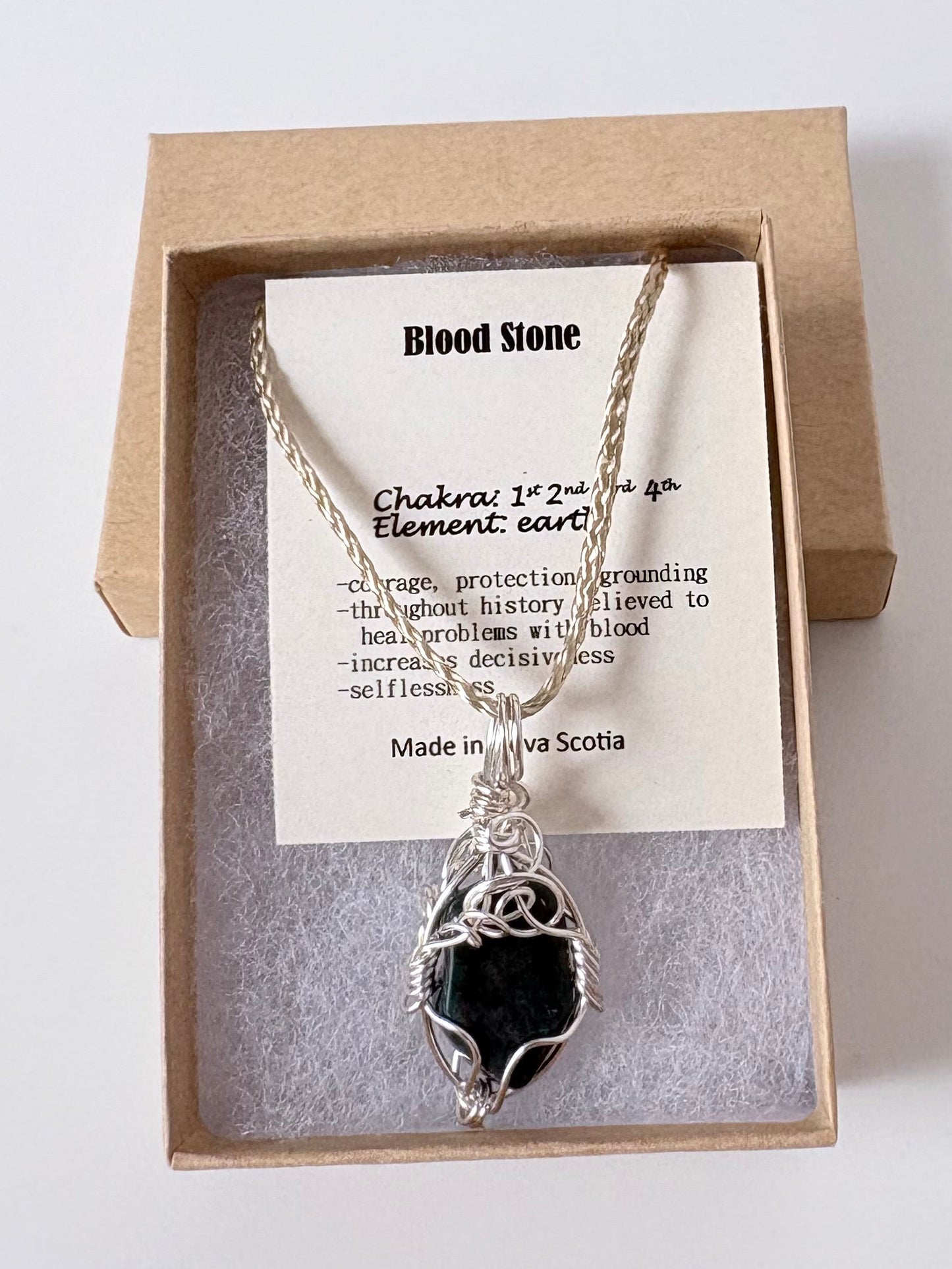 Necklace with a silver wire wrapped bloodstone pendant on an information card with text explaining the folklore and metaphysical properties placed in a brown paper kraft jewelry box for gift giving all on a white surface.