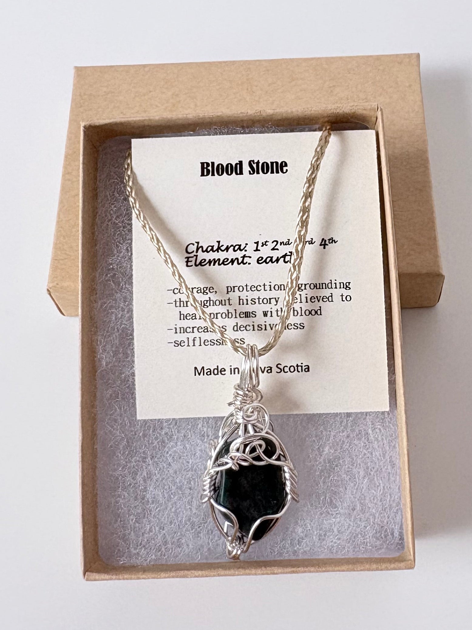 Necklace with a silver wire wrapped bloodstone pendant on an information card with text explaining the folklore and metaphysical properties placed in a brown paper kraft jewelry box for gift giving all on a white surface.