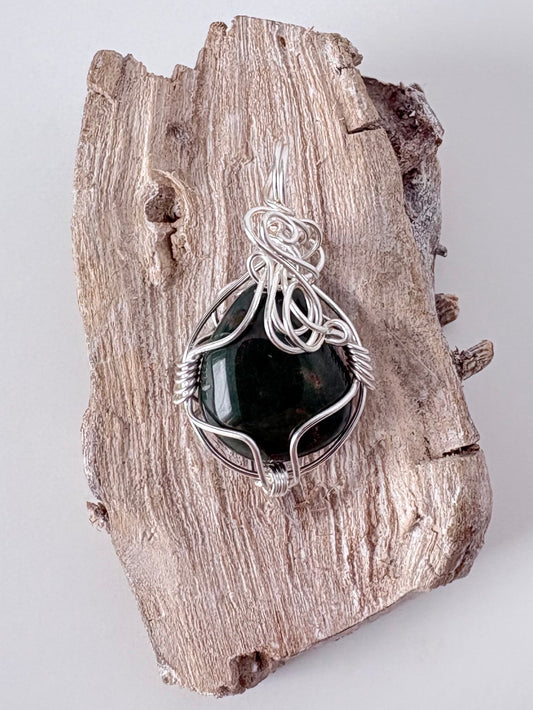 Silver wire-wrapped bloodstone pendant  on a piece of driftwood against a light background