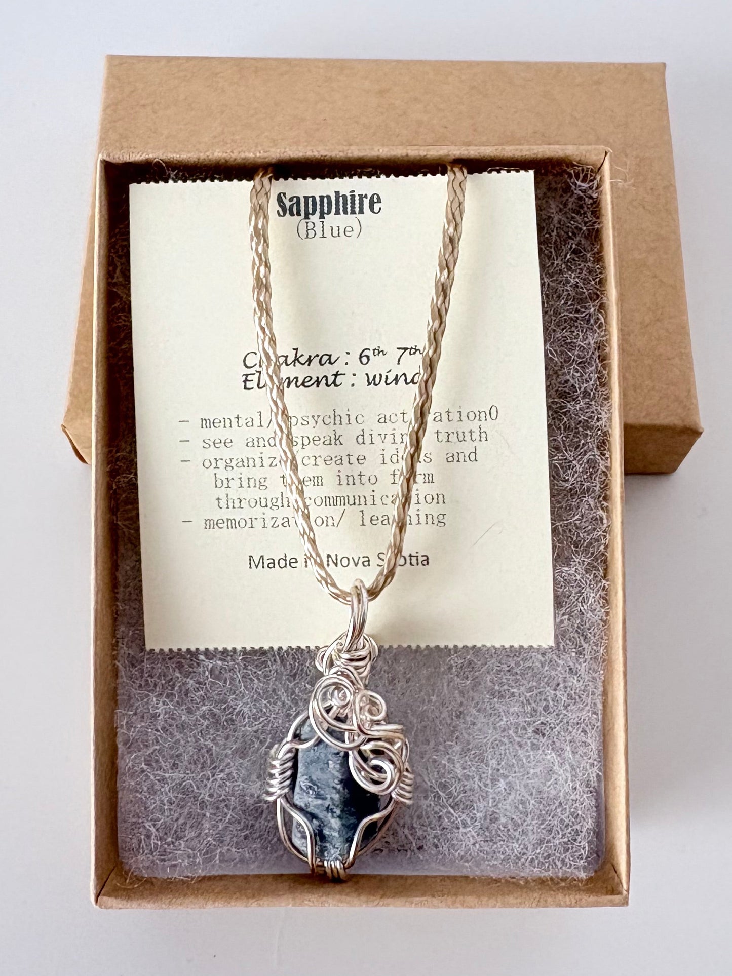 Necklace with a silver wire wrapped blue sapphire pendant on an information card with text explaining the folklore and metaphysical properties placed in a brown paper kraft jewelry box for gift giving all on a white surface.