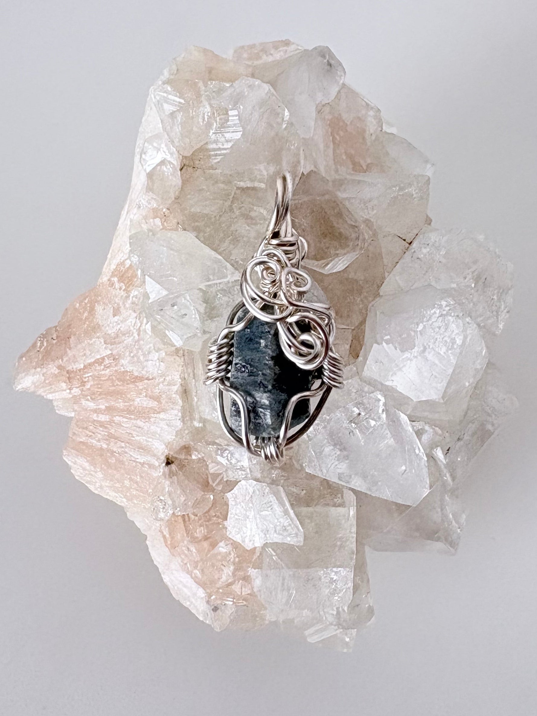 silver wire wrapped blue sapphire pendant displayed on a clear apophyllite cluster all on a white surface.