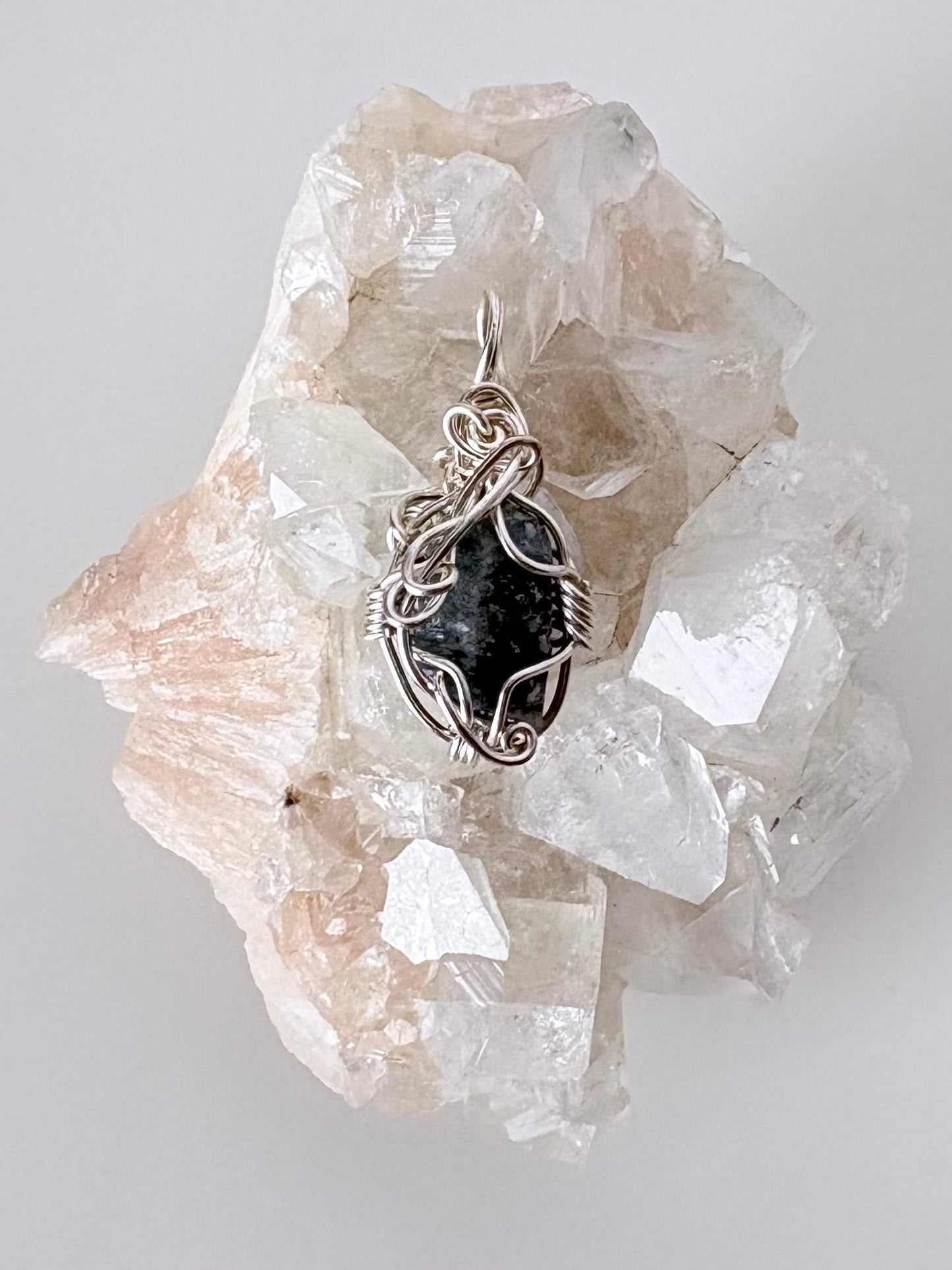 silver wire wrapped blue sapphire pendant displayed on a clear apophyllite cluster all on a white surface.