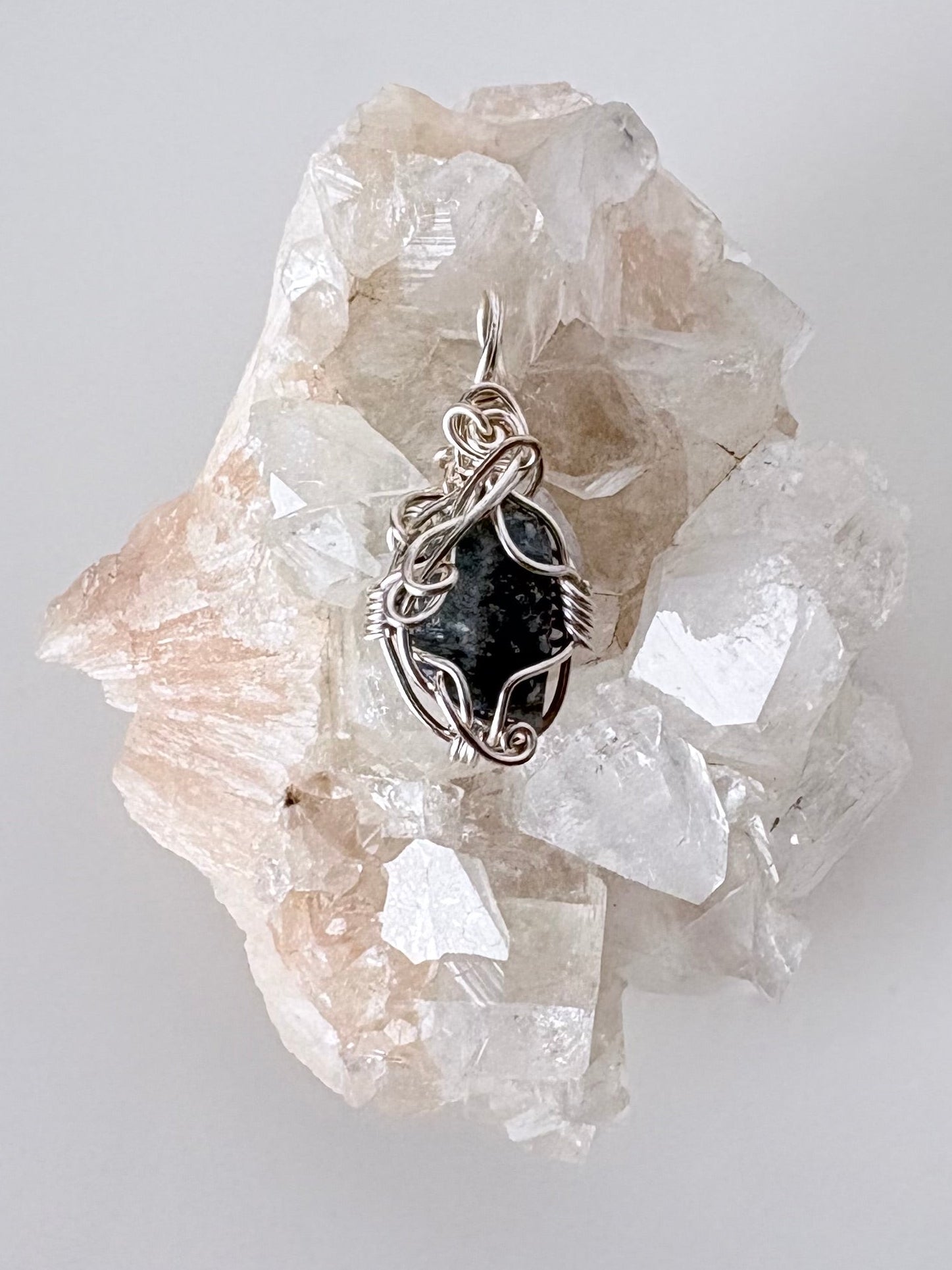 silver wire wrapped blue sapphire pendant displayed on a clear apophyllite cluster all on a white surface.