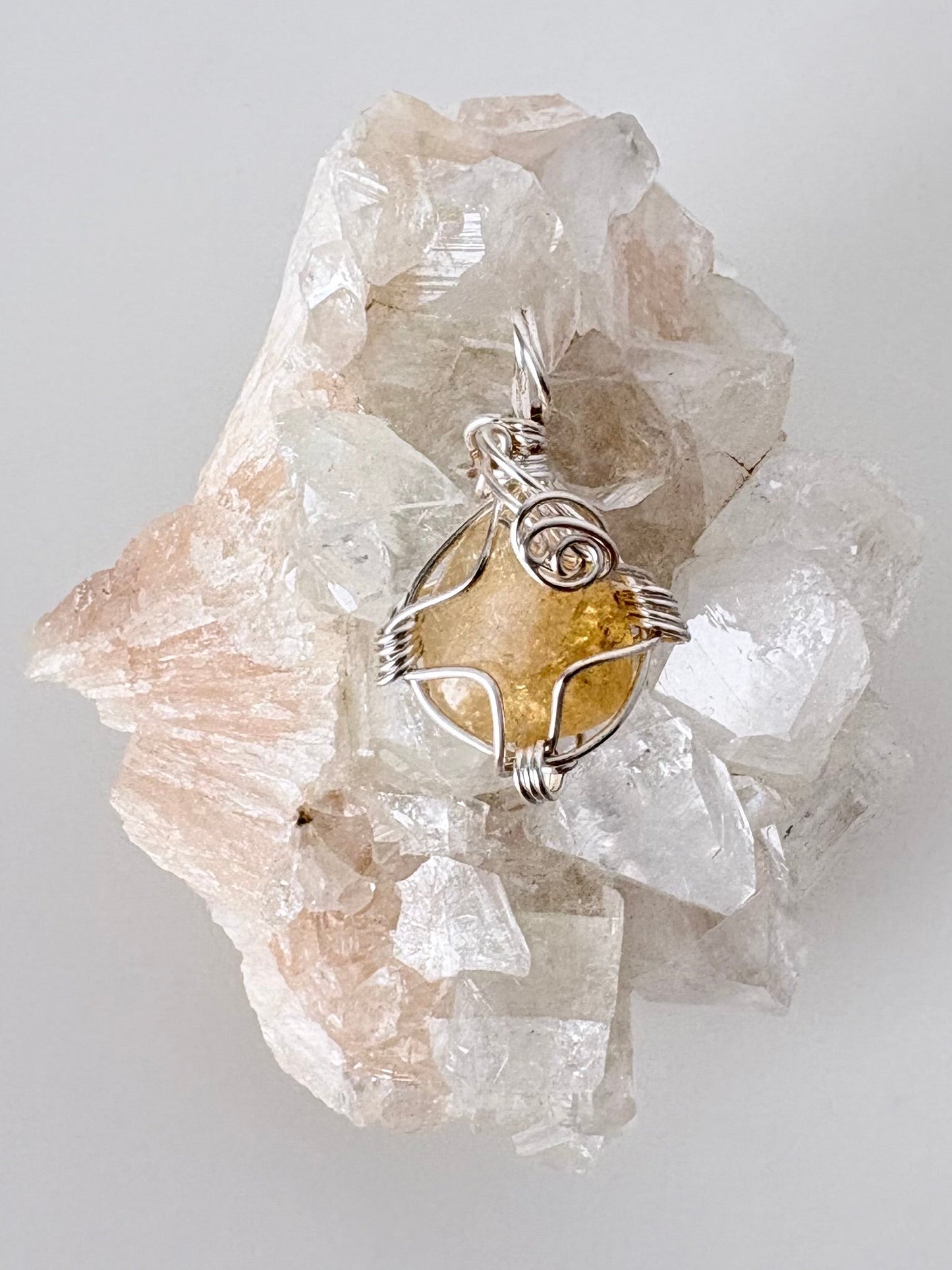 silver wire wrapped citrine pendant displayed on a clear apophyllite cluster all on a white surface.