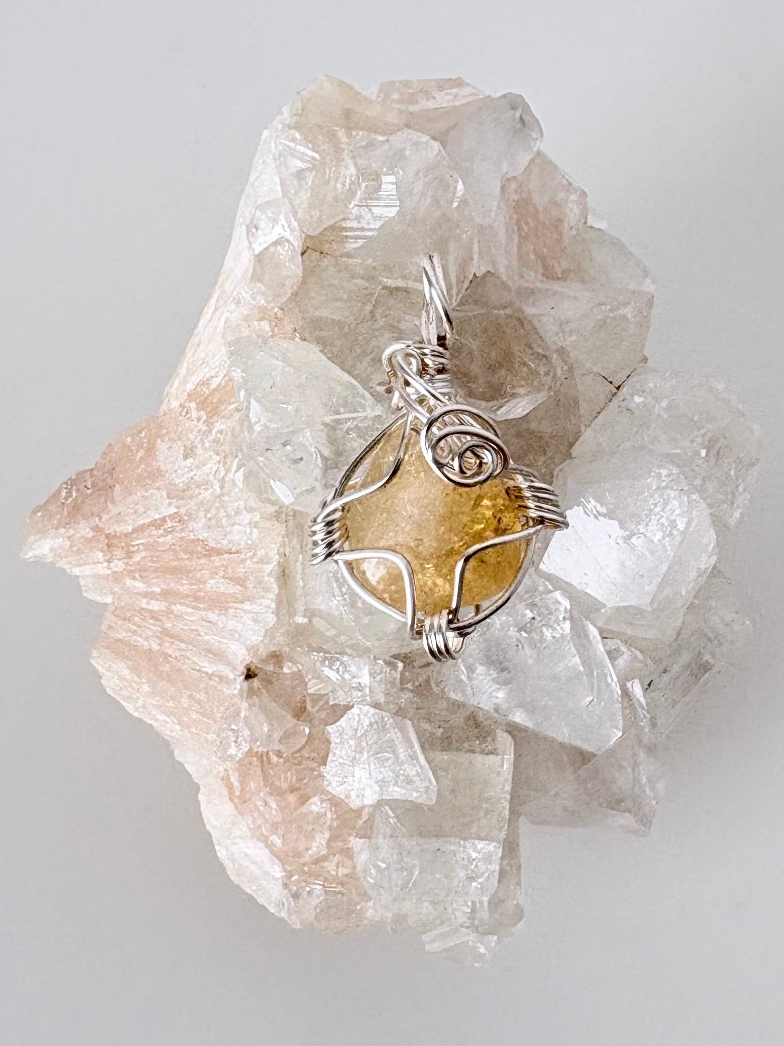 silver wire wrapped citrine pendant displayed on a clear apophyllite cluster all on a white surface.