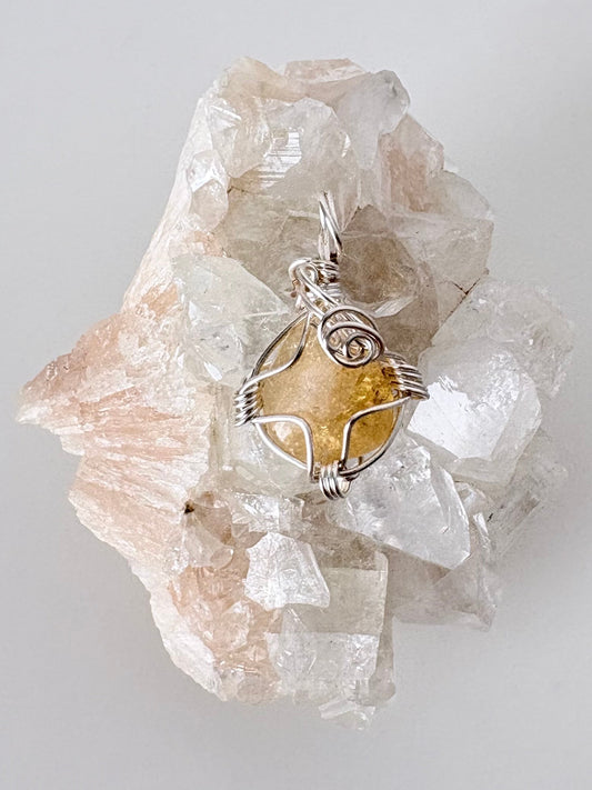 silver wire wrapped citrine pendant displayed on a clear apophyllite cluster all on a white surface.