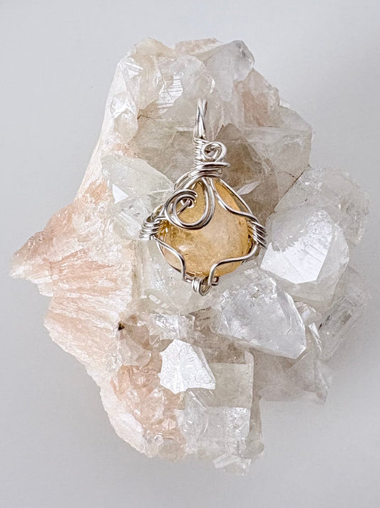 silver wire wrapped citrine pendant displayed on a clear apophyllite cluster all on a white surface.