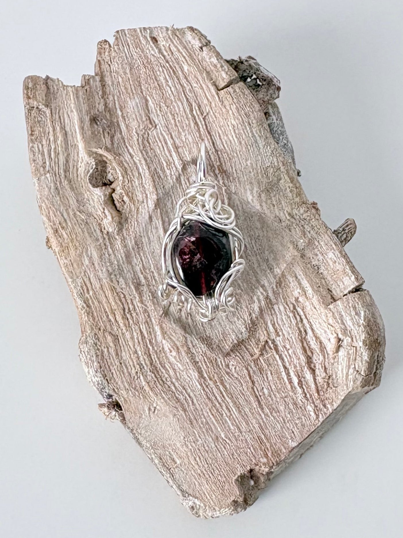 Silver wire-wrapped garnet pendant  on a piece of driftwood against a light background