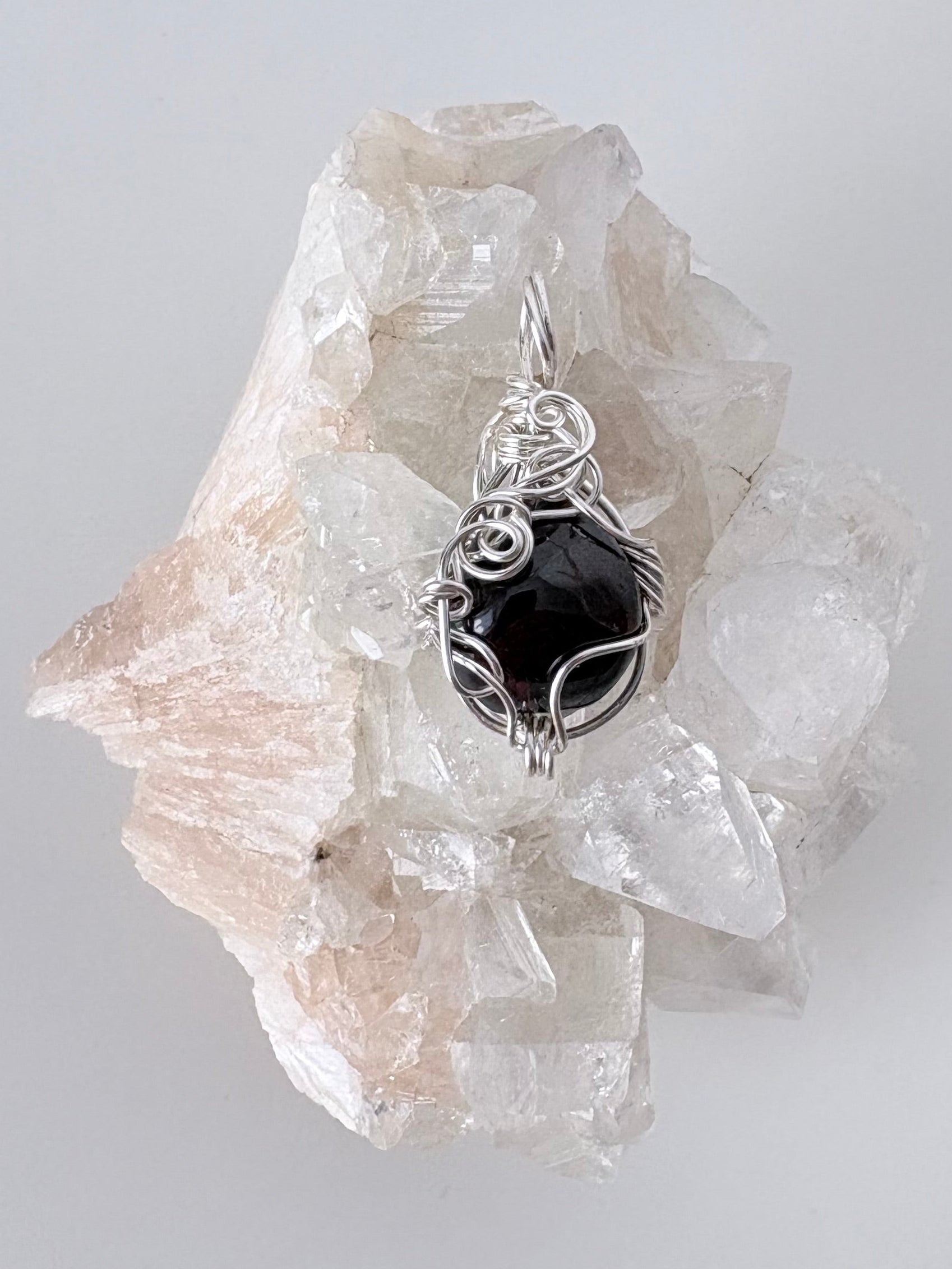 silver wire wrapped garnet pendant displayed on a clear apophyllite cluster all on a white surface.