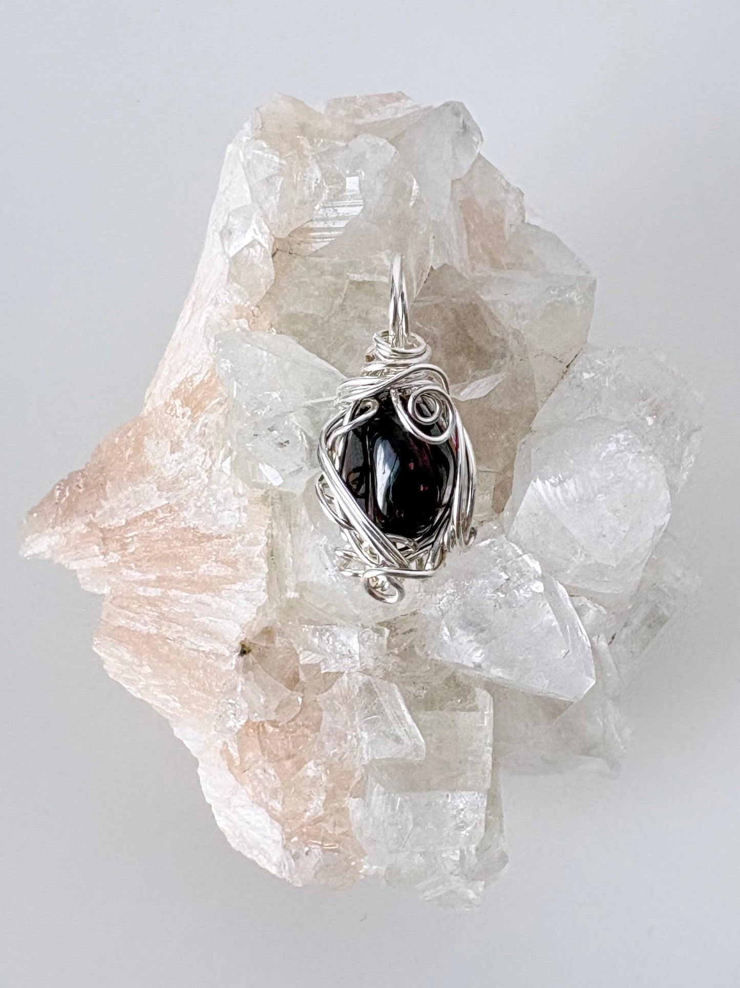 silver wire wrapped garnet pendant displayed on a clear apophyllite cluster all on a white surface.