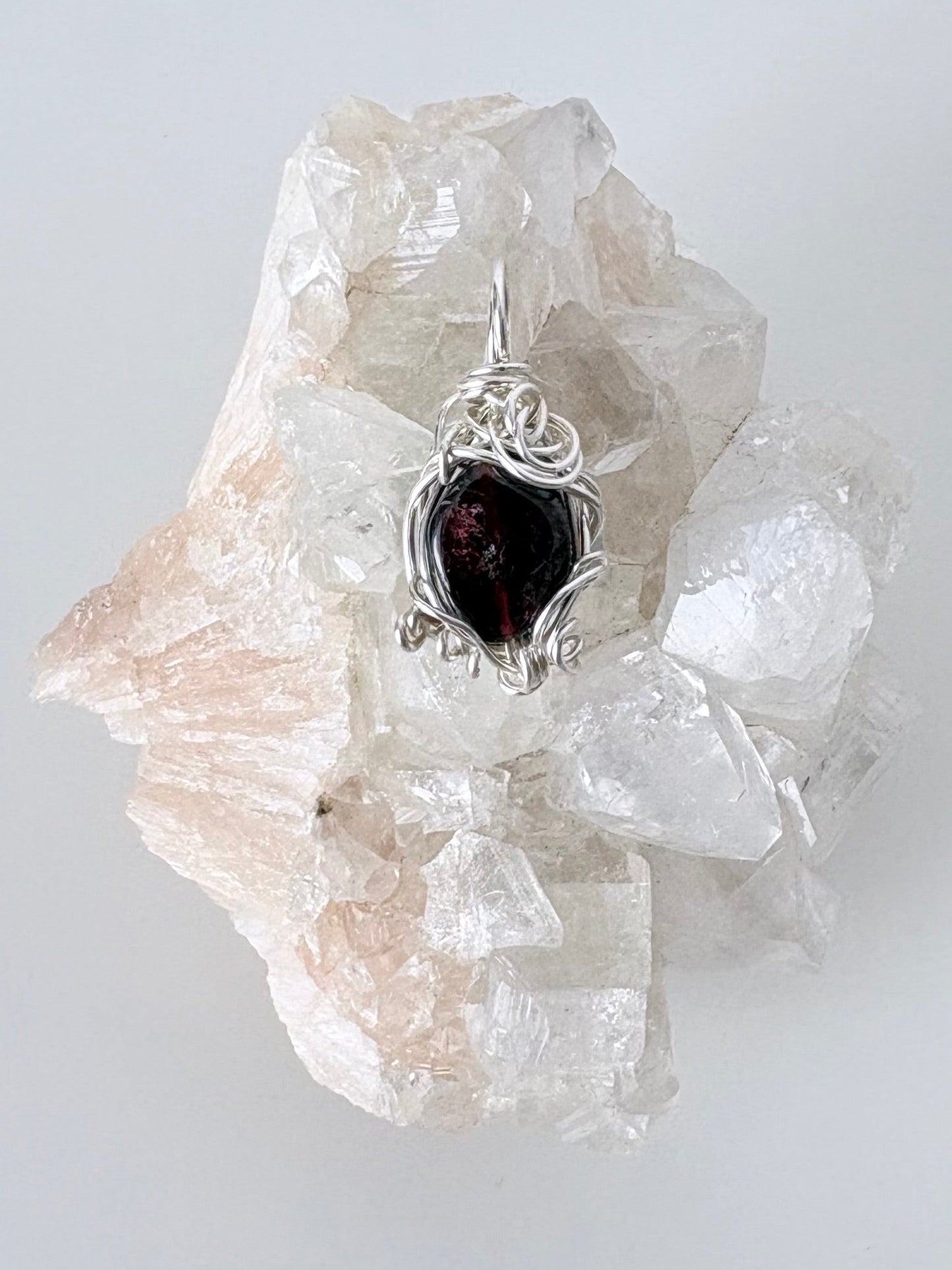 silver wire wrapped garnet pendant displayed on a clear apophyllite cluster all on a white surface.