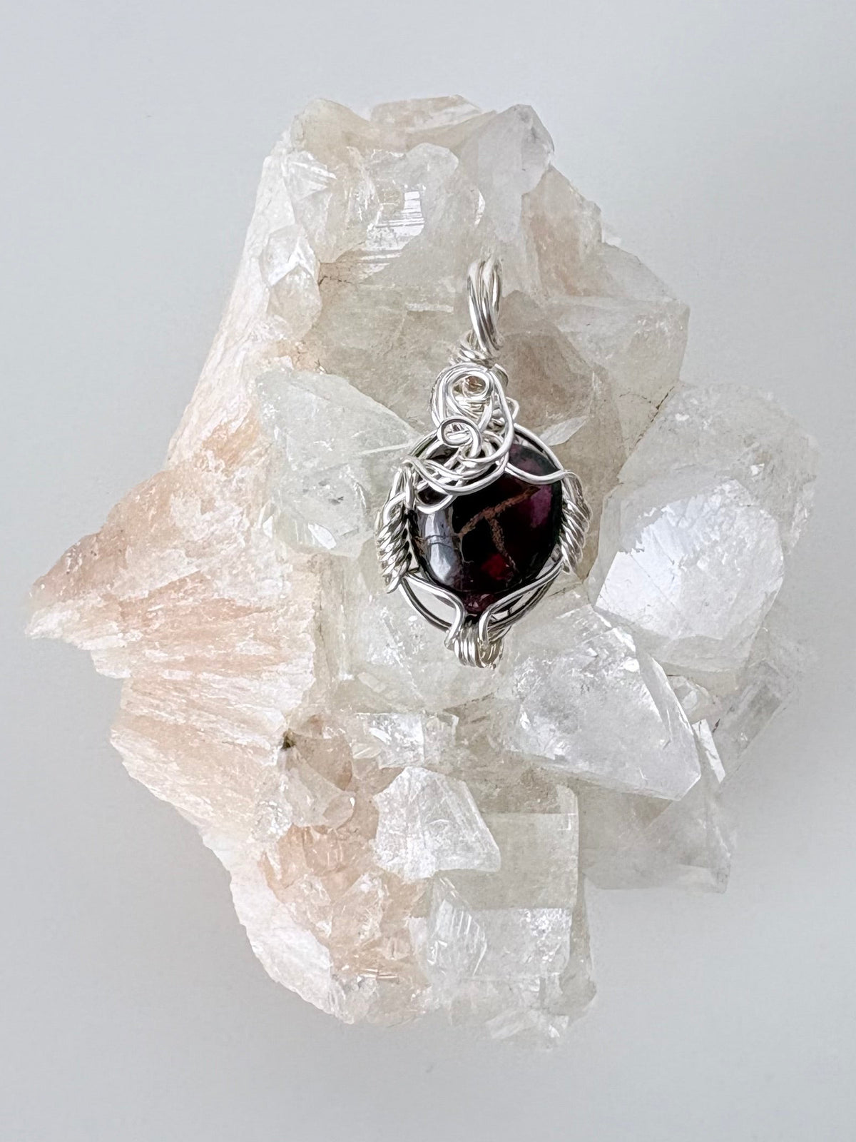 silver wire wrapped garnet pendant displayed on a clear apophyllite cluster all on a white surface.