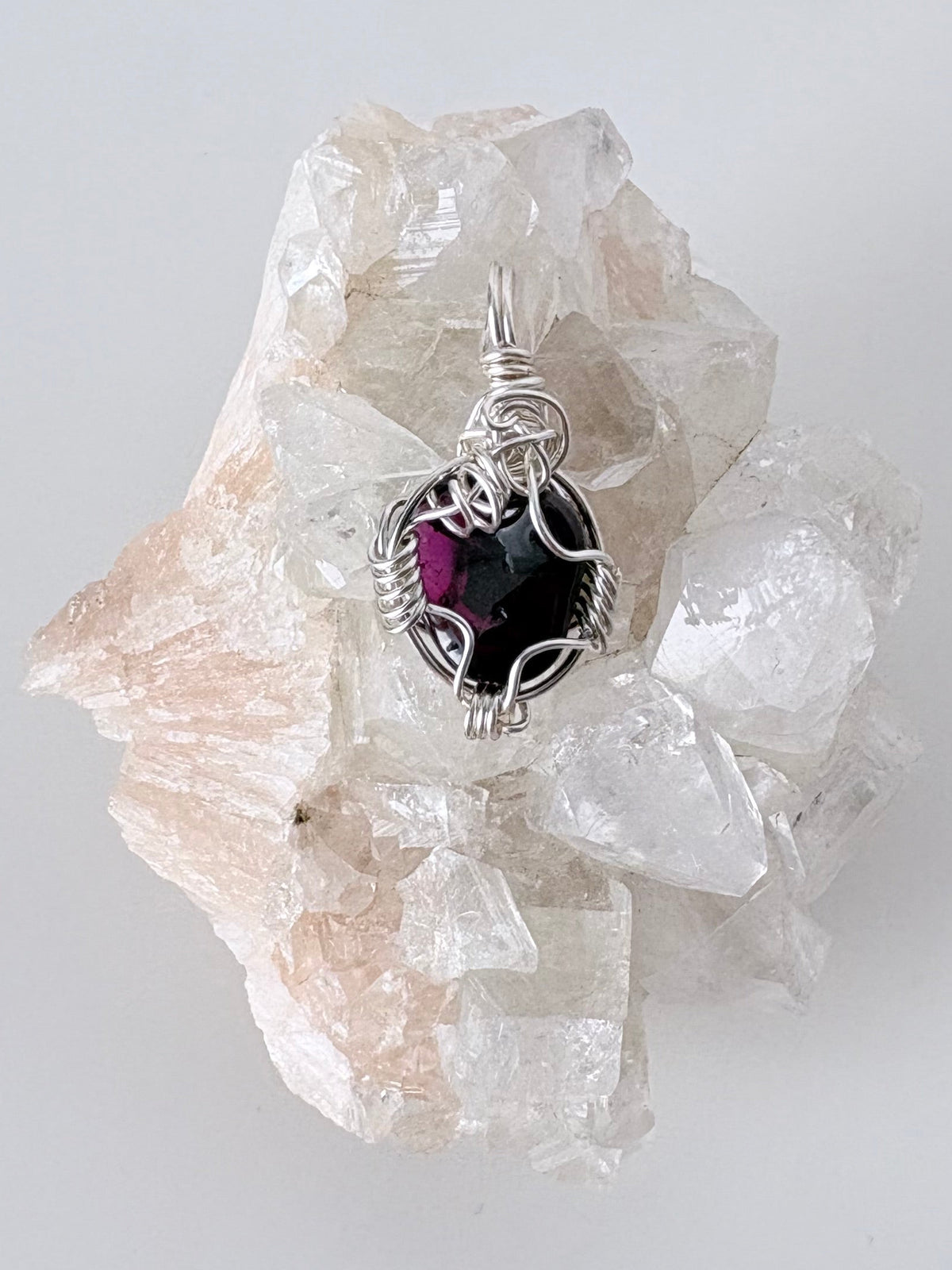 silver wire wrapped garnet pendant displayed on a clear apophyllite cluster all on a white surface.