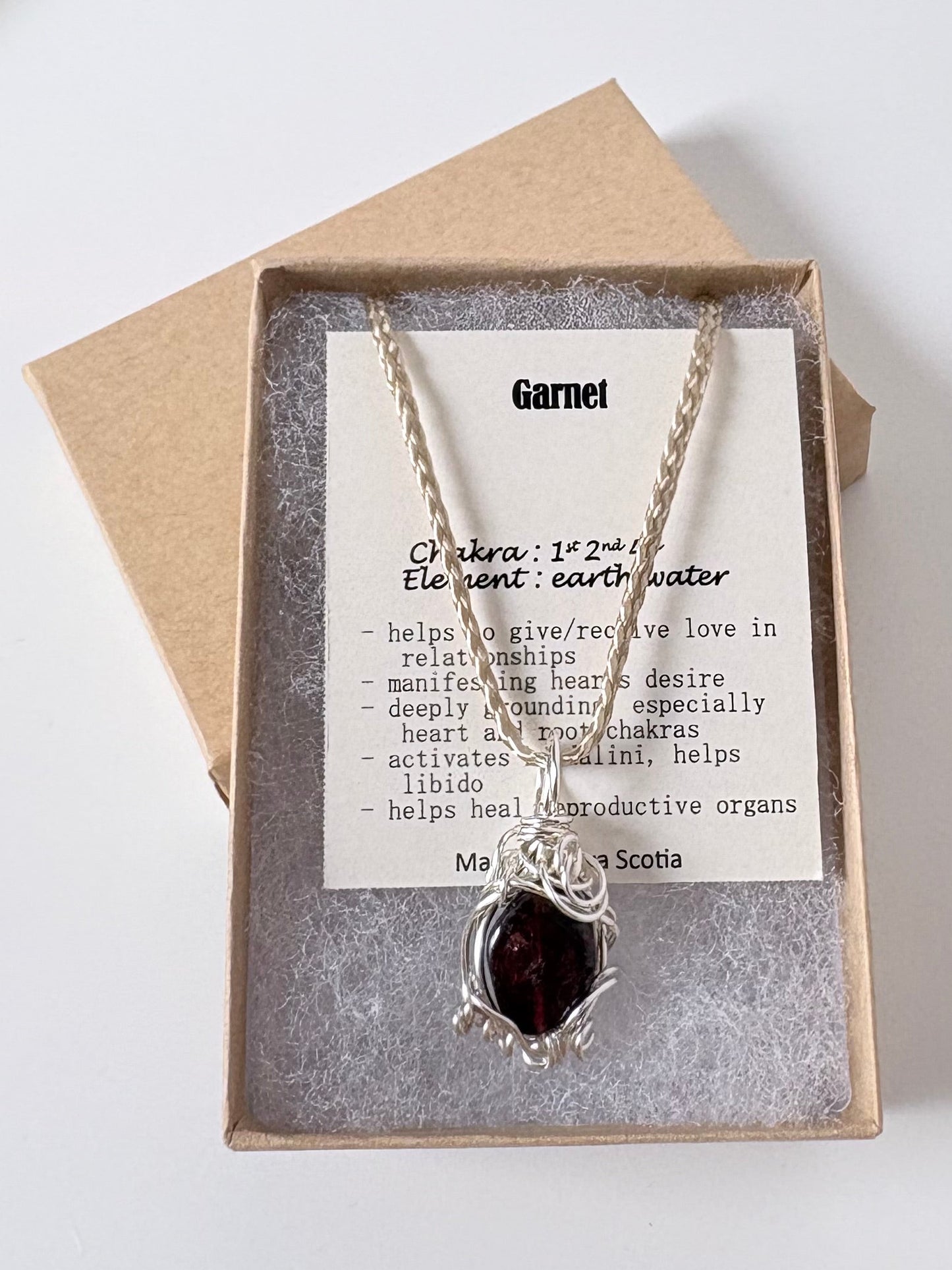 Necklace with a silver wire wrapped garnet pendant on an information card with text explaining the folklore and metaphysical properties placed in a brown paper kraft jewelry box for gift giving all on a white surface.