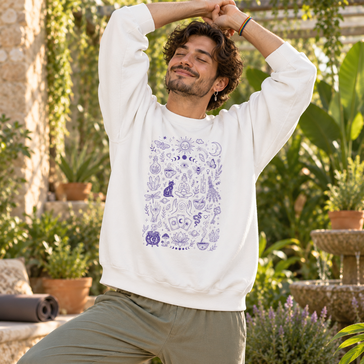 A man wearing a white crewneck sweatshirt with a purple celestial pattern, posing outdoors in a garden setting.