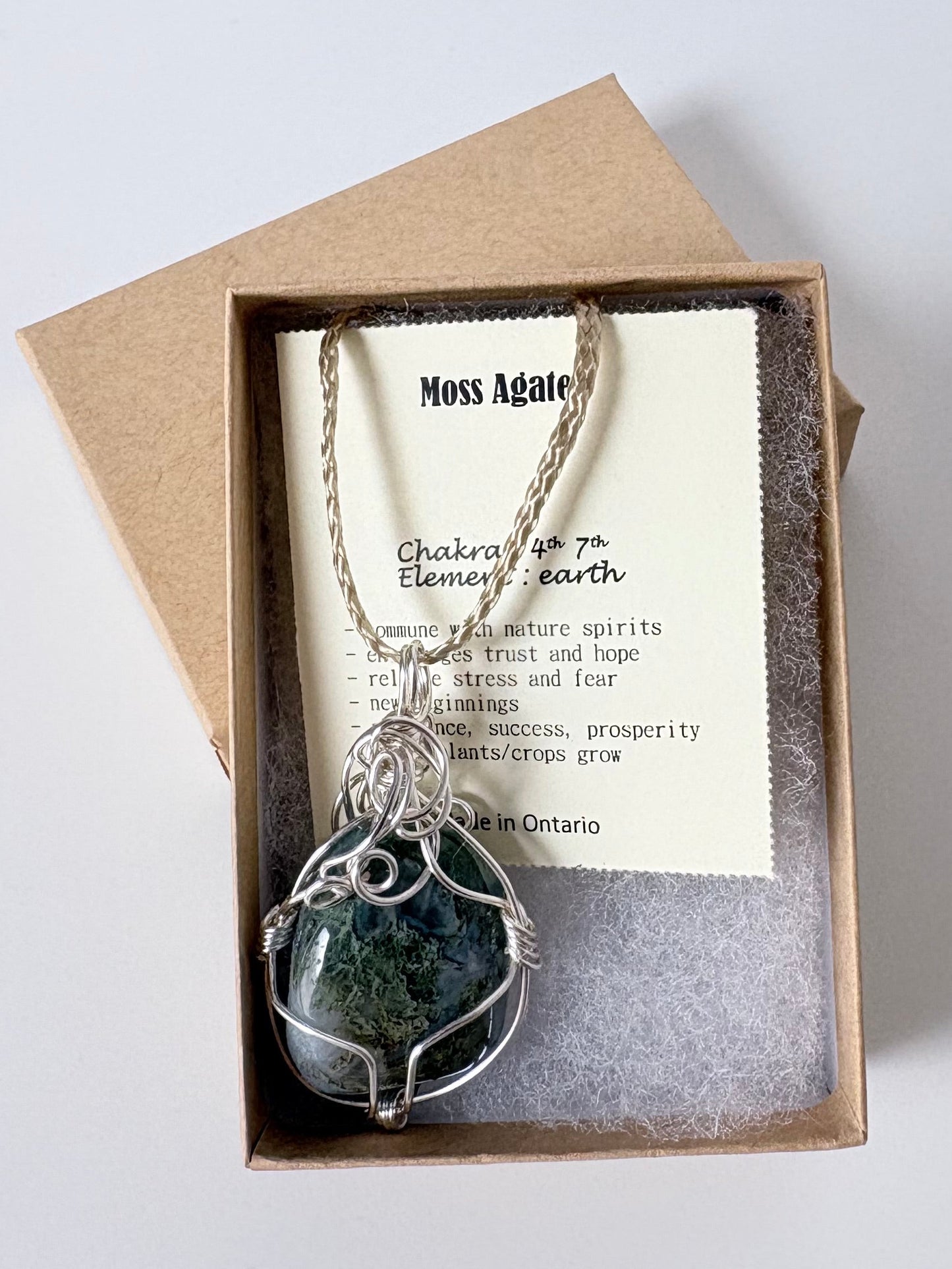 Necklace with a silver wire wrapped moss agate pendant on an information card with text explaining the folklore and metaphysical properties placed in a brown paper kraft jewelry box for gift giving all on a white surface.