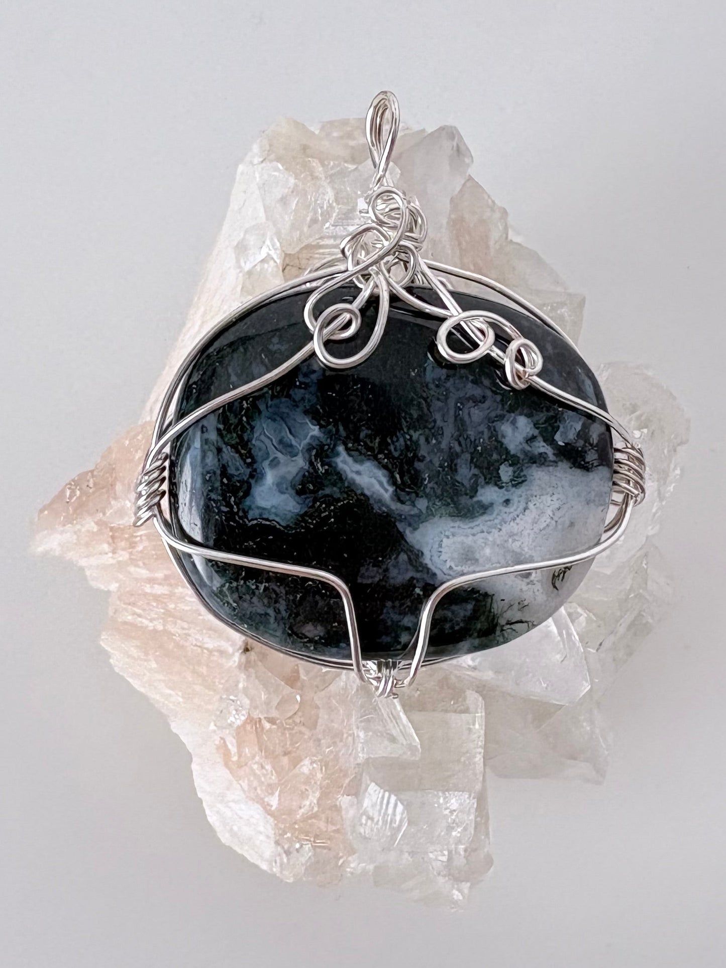 silver wire wrapped moss agate pendant displayed on a clear apophyllite cluster all on a white surface.