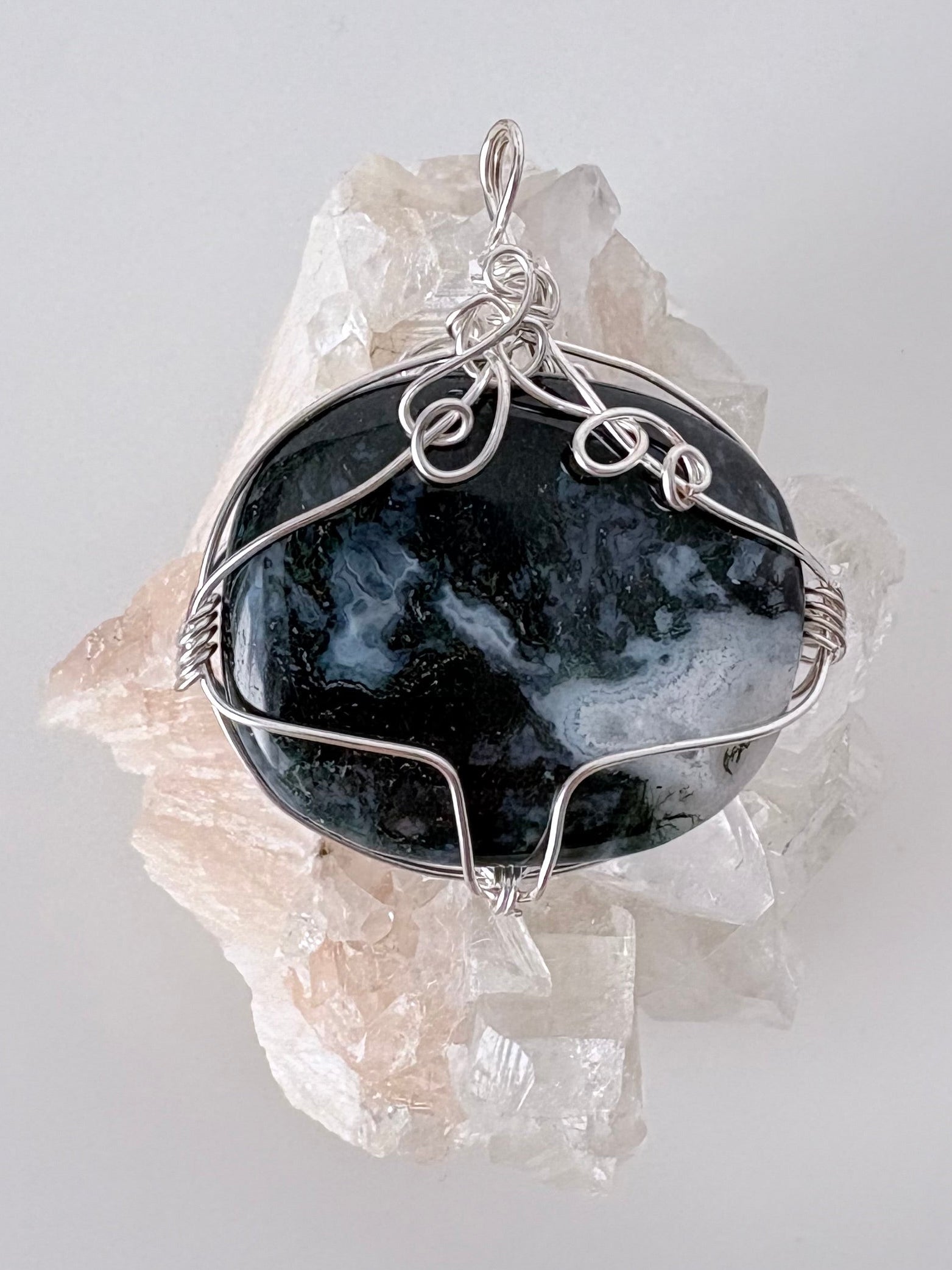 silver wire wrapped moss agate pendant displayed on a clear apophyllite cluster all on a white surface.