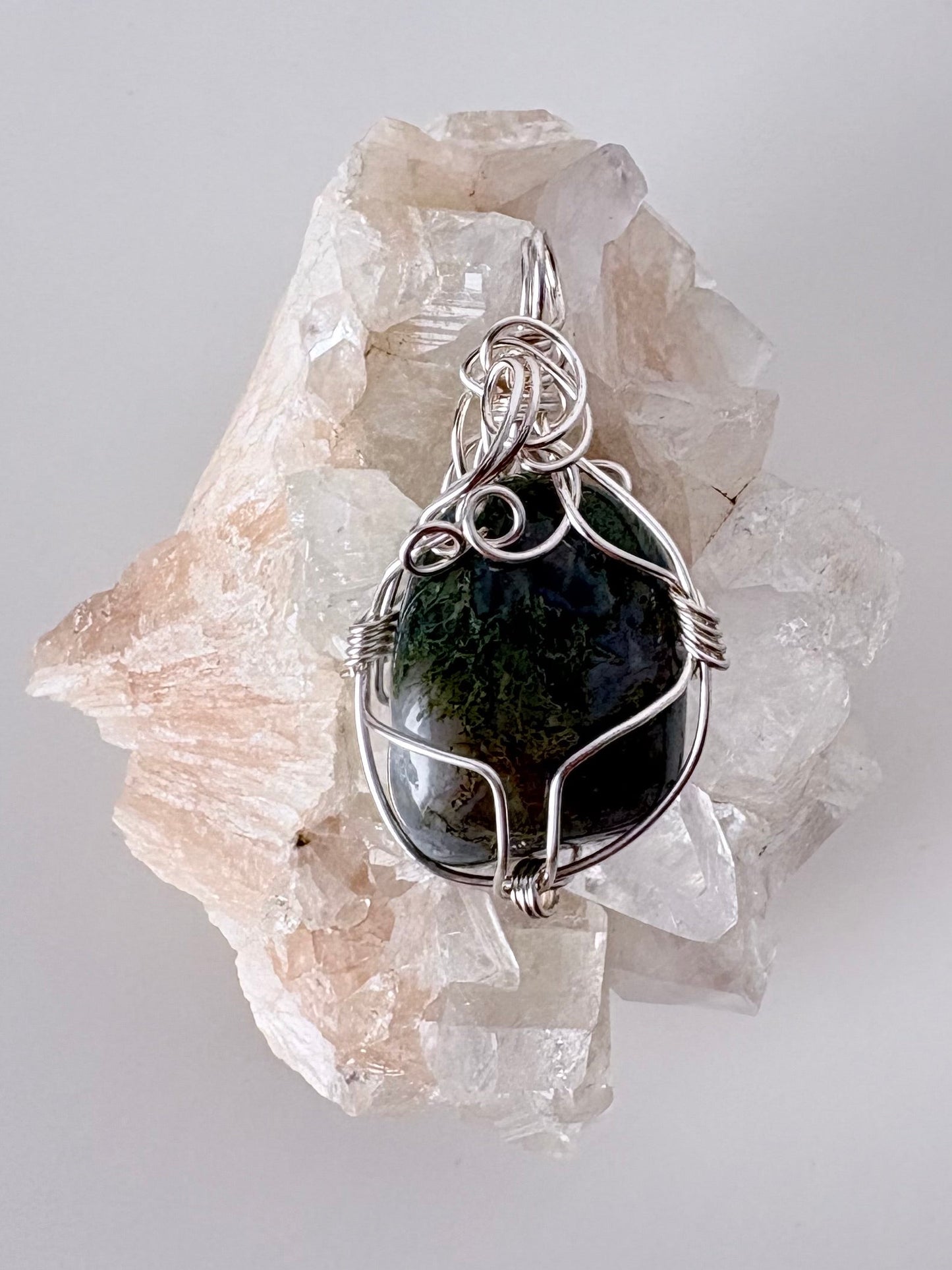 silver wire wrapped moss agate pendant displayed on a clear apophyllite cluster all on a white surface.