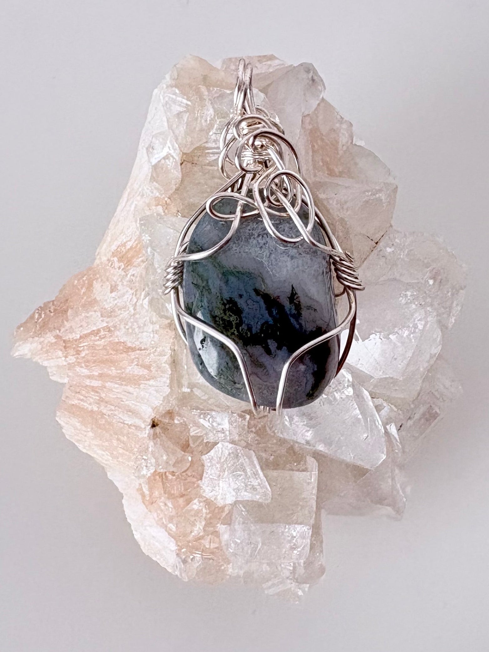 silver wire wrapped moss agate pendant displayed on a clear apophyllite cluster all on a white surface.