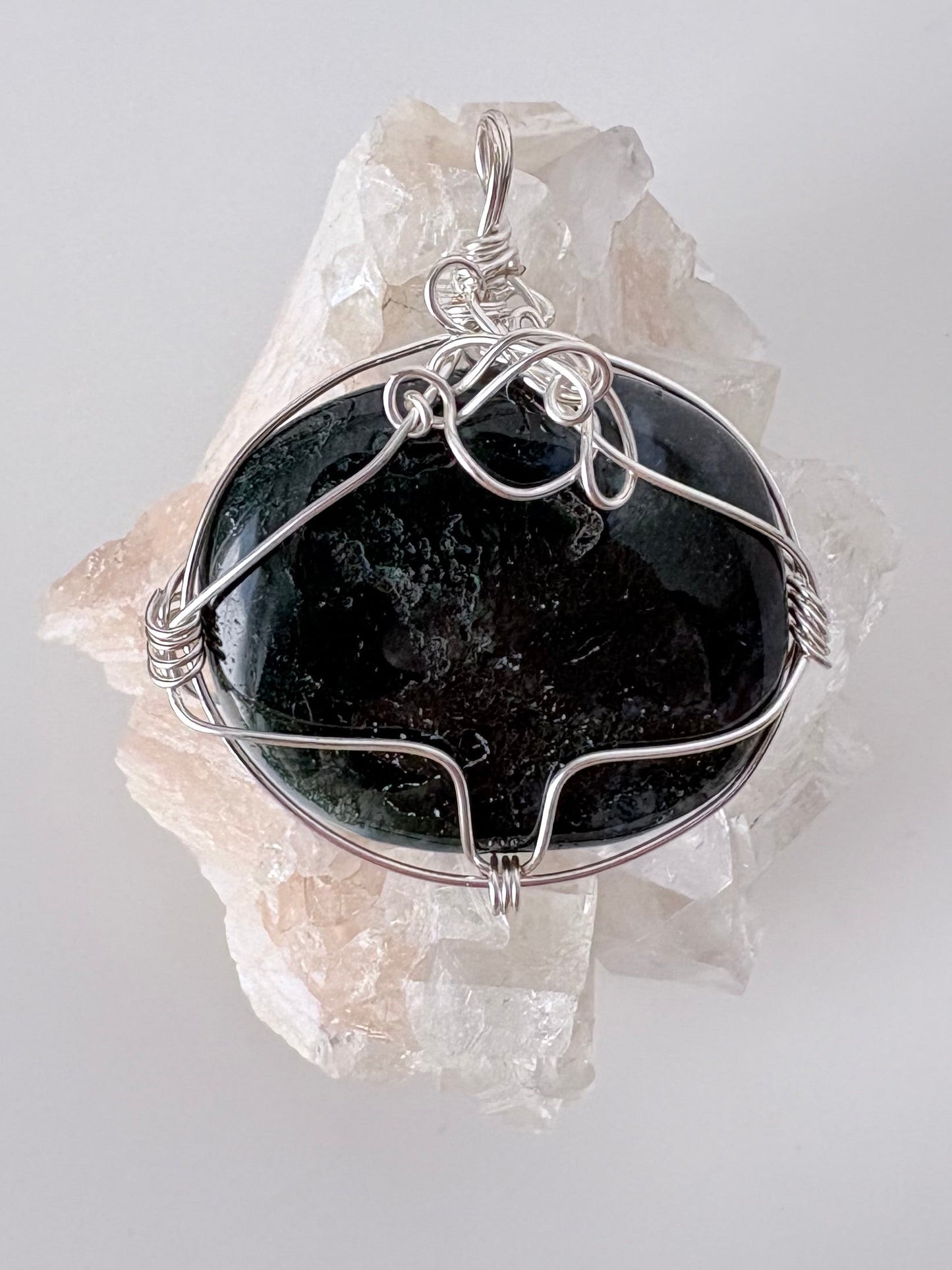 silver wire wrapped moss agate pendant displayed on a clear apophyllite cluster all on a white surface.