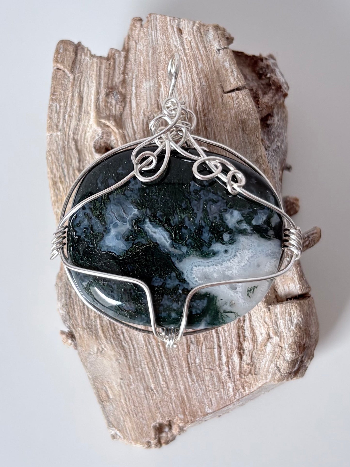 Silver wire-wrapped moss agate pendant  on a piece of driftwood against a light background