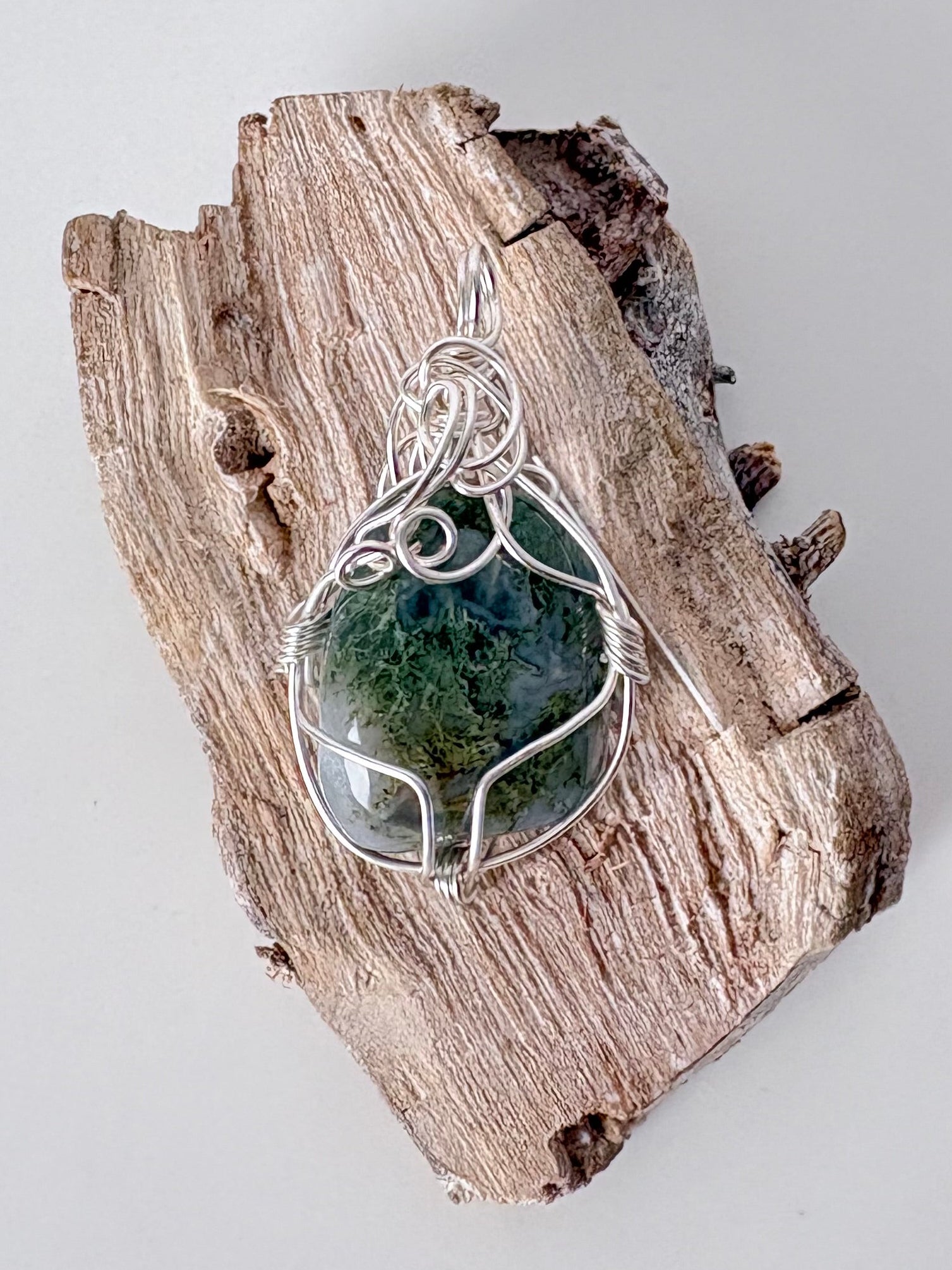 Silver wire-wrapped moss agate pendant  on a piece of driftwood against a light background