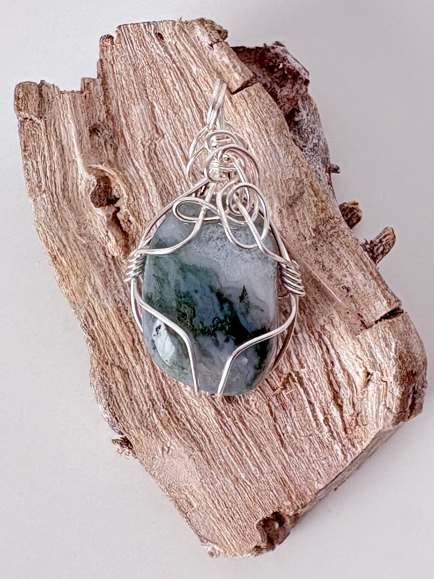 Silver wire-wrapped moss agate pendant  on a piece of driftwood against a light background