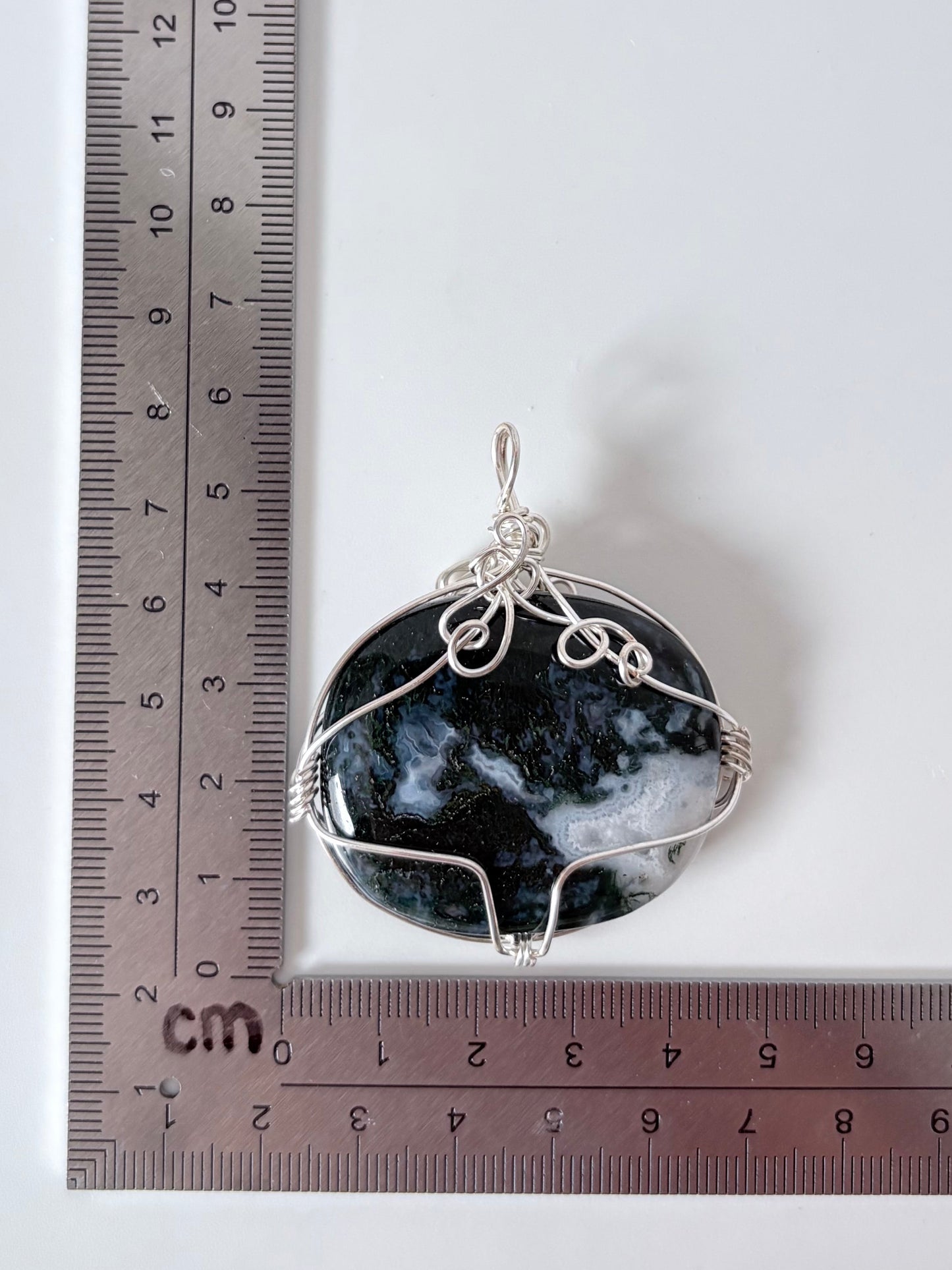 silver wire wrapped moss agate pendant next to an "L" shaped ruler for scale all against a white background.