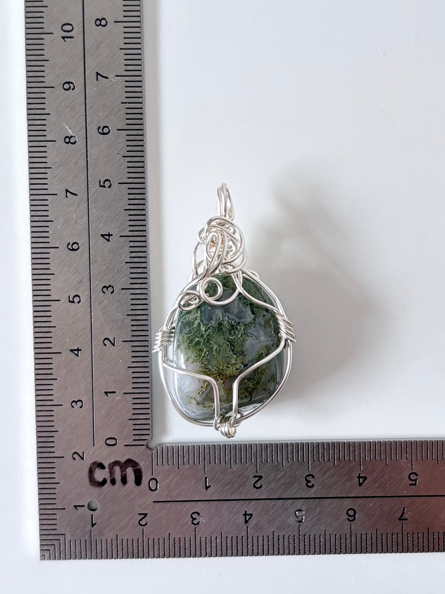 silver wire wrapped moss agate pendant next to an "L" shaped ruler for scale all against a white background.