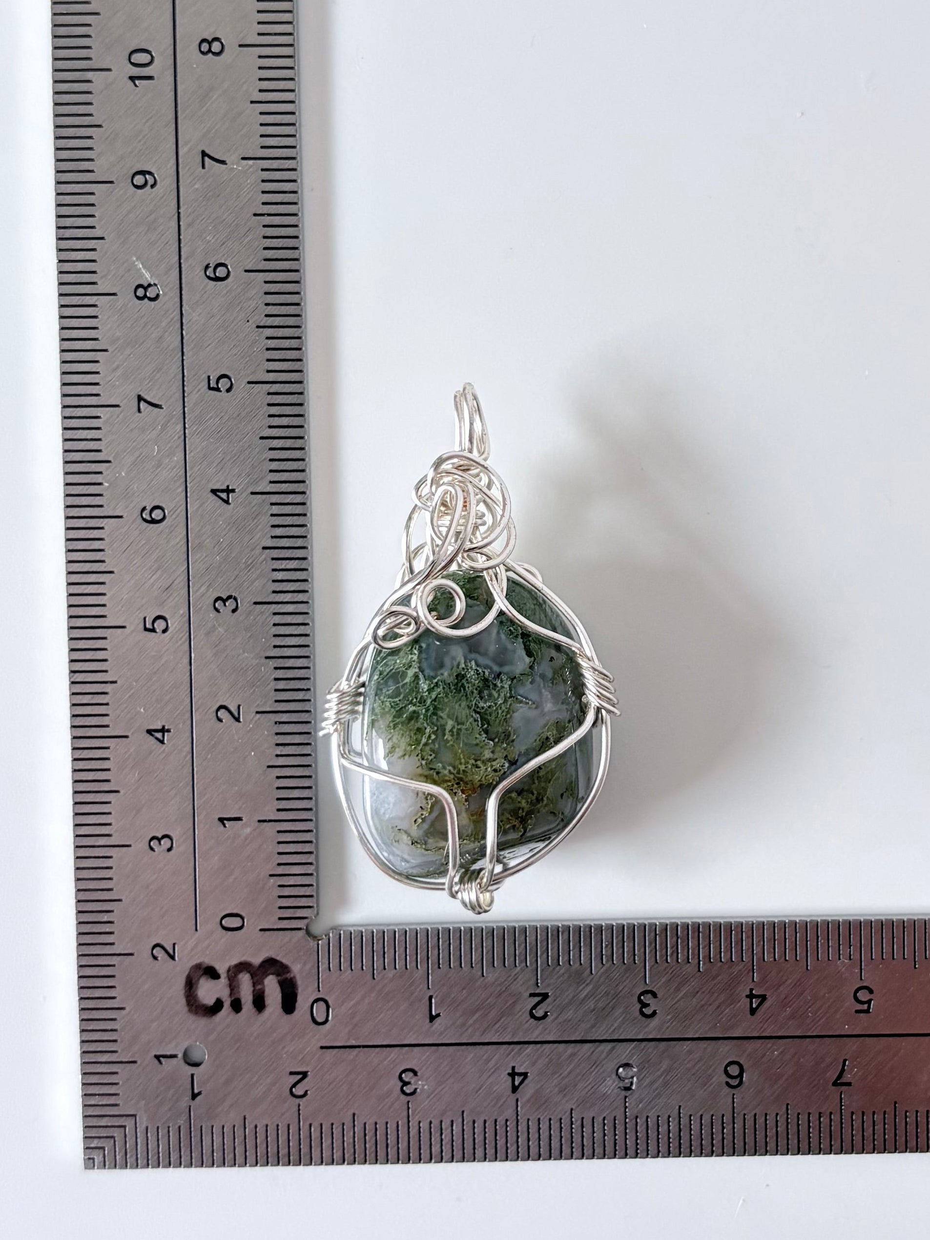 silver wire wrapped moss agate pendant next to an "L" shaped ruler for scale all against a white background.