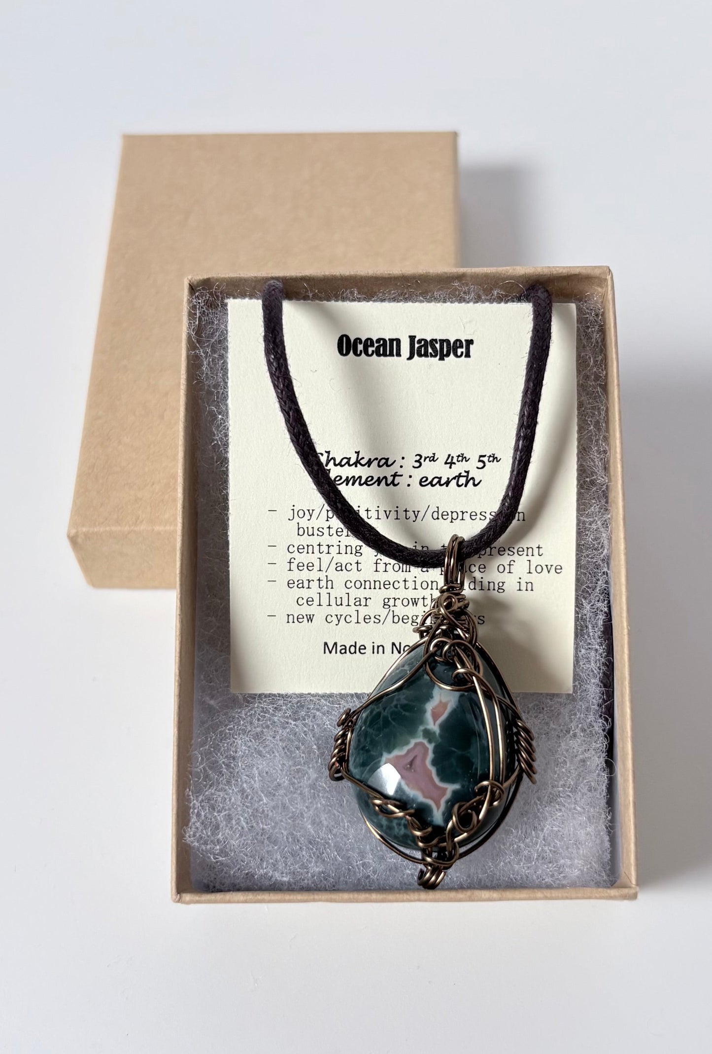 Necklace with a silver wire wrapped ocean jasper pendant on an information card with text explaining the folklore and metaphysical properties placed in a brown paper kraft jewelry box for gift giving all on a white surface.