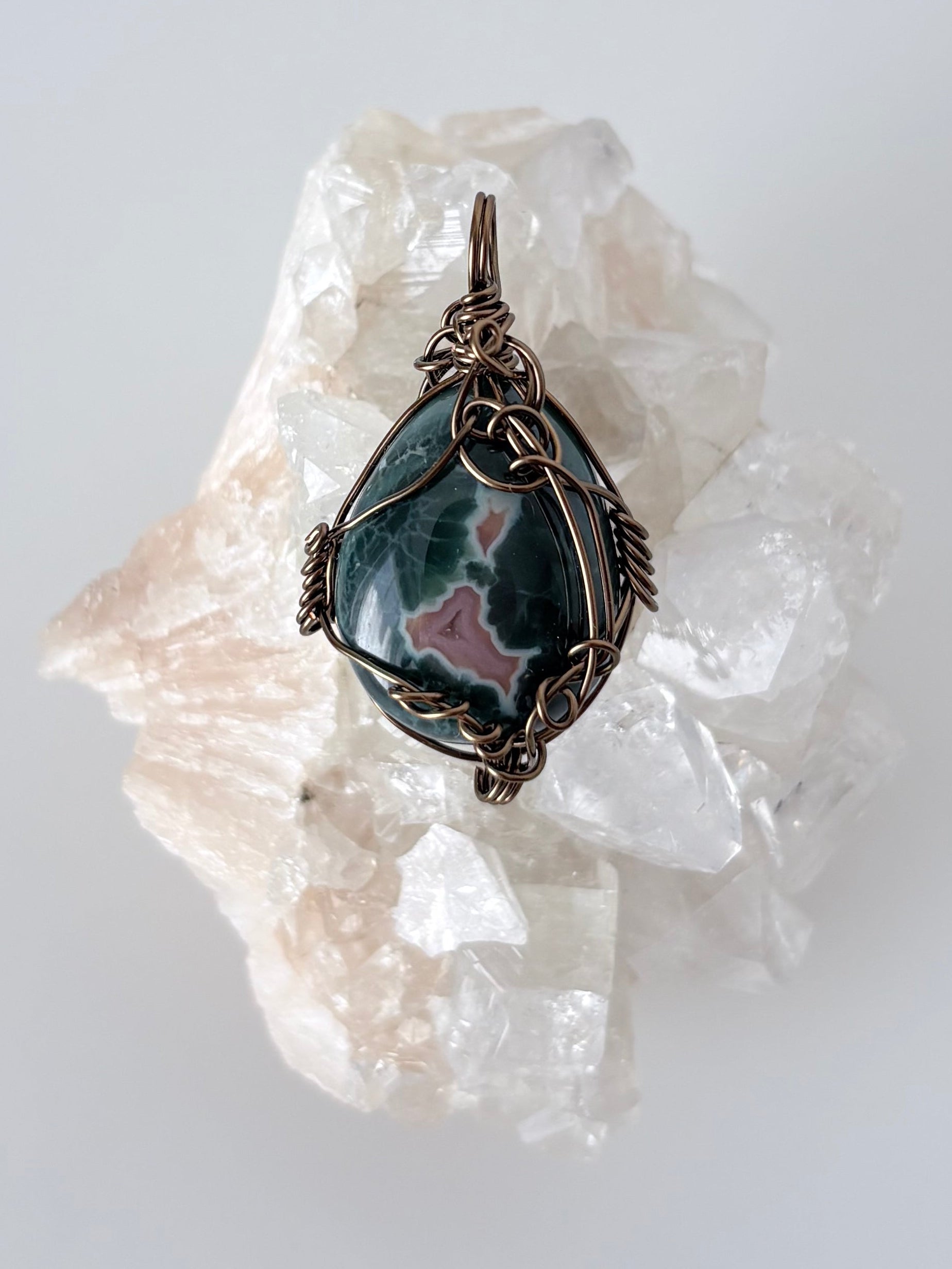 silver wire wrapped ocean jasper pendant displayed on a clear apophyllite cluster all on a white surface.