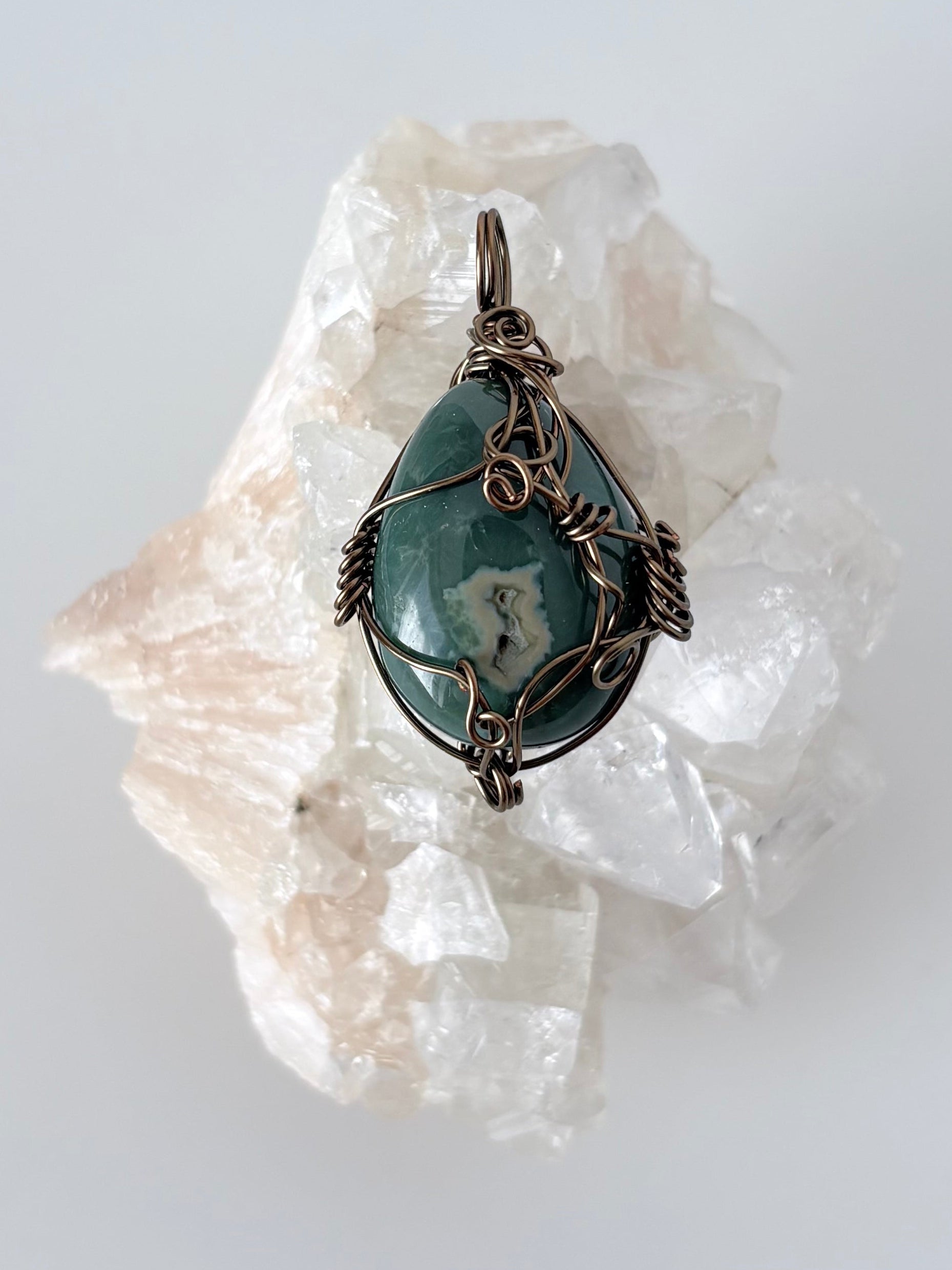 silver wire wrapped ocean jasper pendant displayed on a clear apophyllite cluster all on a white surface.
