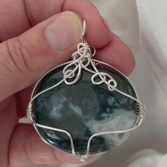 hand holding and moving the silver wire wrapped moss agate pendant around so all the sides of it are shown. done against a white background.